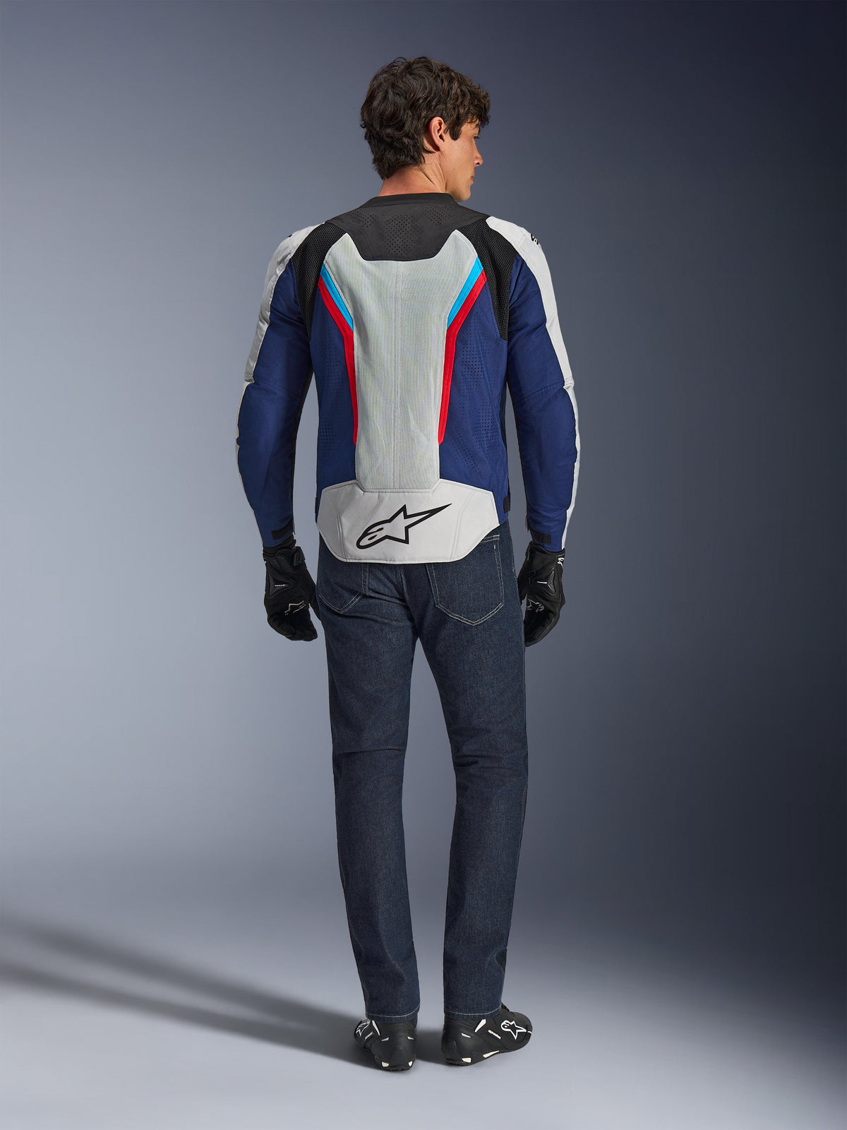 Alpinestars AST V3 Air Jacket, rear view, Silver Deep Blue and Bright Red, featuring a large mesh ventilation panel on the back with the Alpinestars logo, worn with dark blue jeans and black gloves, sport-fit warm-weather motorcycle gear