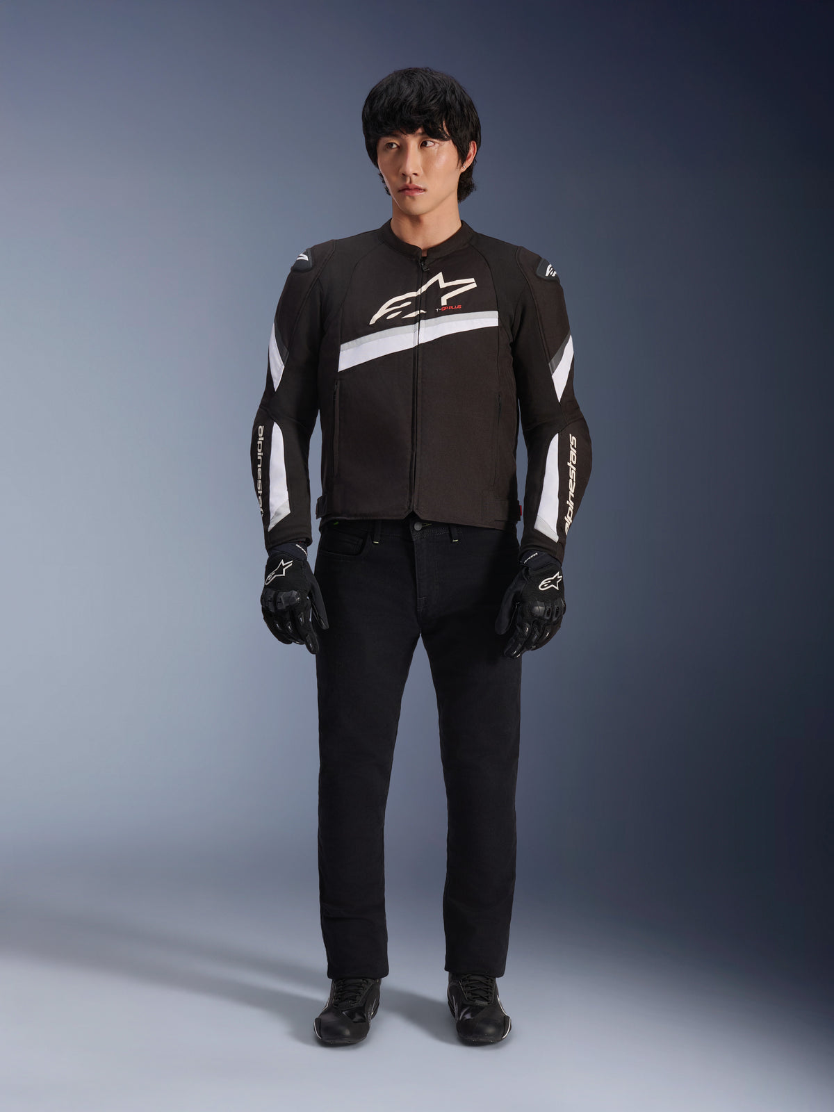 Chaqueta Alpinestars T-GP Plus R V4, negra y blanca, chaqueta de moto de textil deportiva, lucida por un modelo con pantalones y guantes negros, con el logotipo blanco de Alpinestars en el pecho y las mangas, diseño elegante inspirado en las carreras