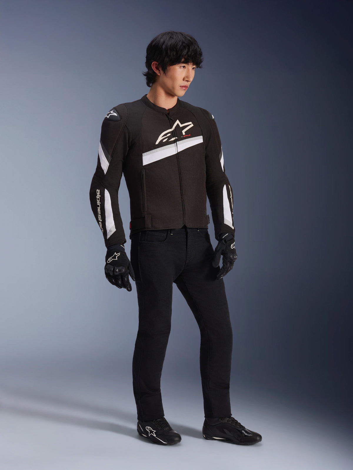 Chaqueta Alpinestars T-GP Plus R V4, chaqueta textil de moto en color negro y blanco usada por un modelo con pantalones y guantes negros, con logotipos blancos de Alpinestars en el pecho y las mangas, diseño deportivo para rendimiento en carretera