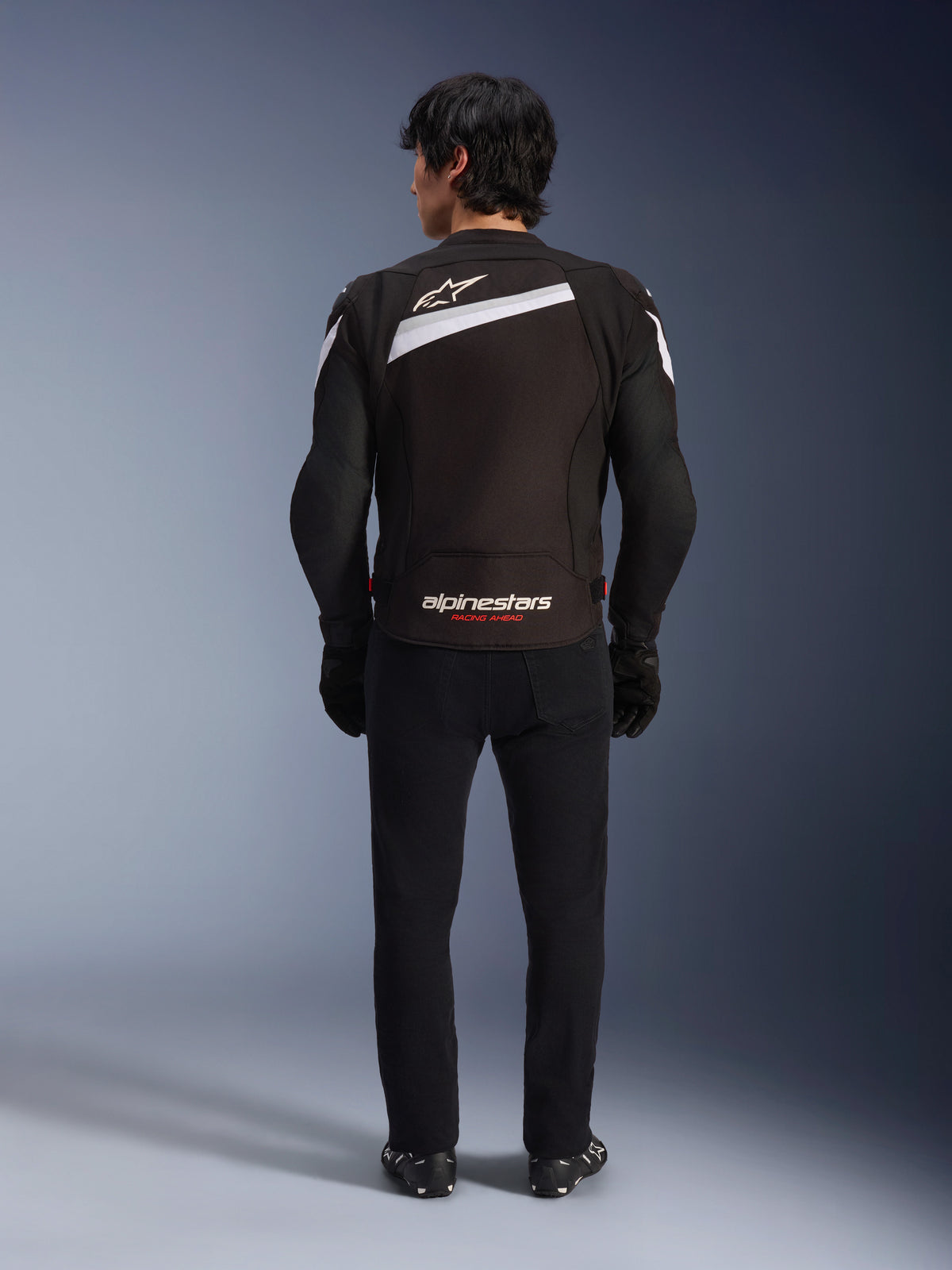 Chaqueta Alpinestars T-GP Plus R V4, chaqueta de moto textil deportiva en negro y blanco, vista trasera con el logotipo blanco de Alpinestars y la inscripción 'RACING AHEAD', combinada con pantalones y botas negros, diseño aerodinámico para una conducción de alto rendimiento.