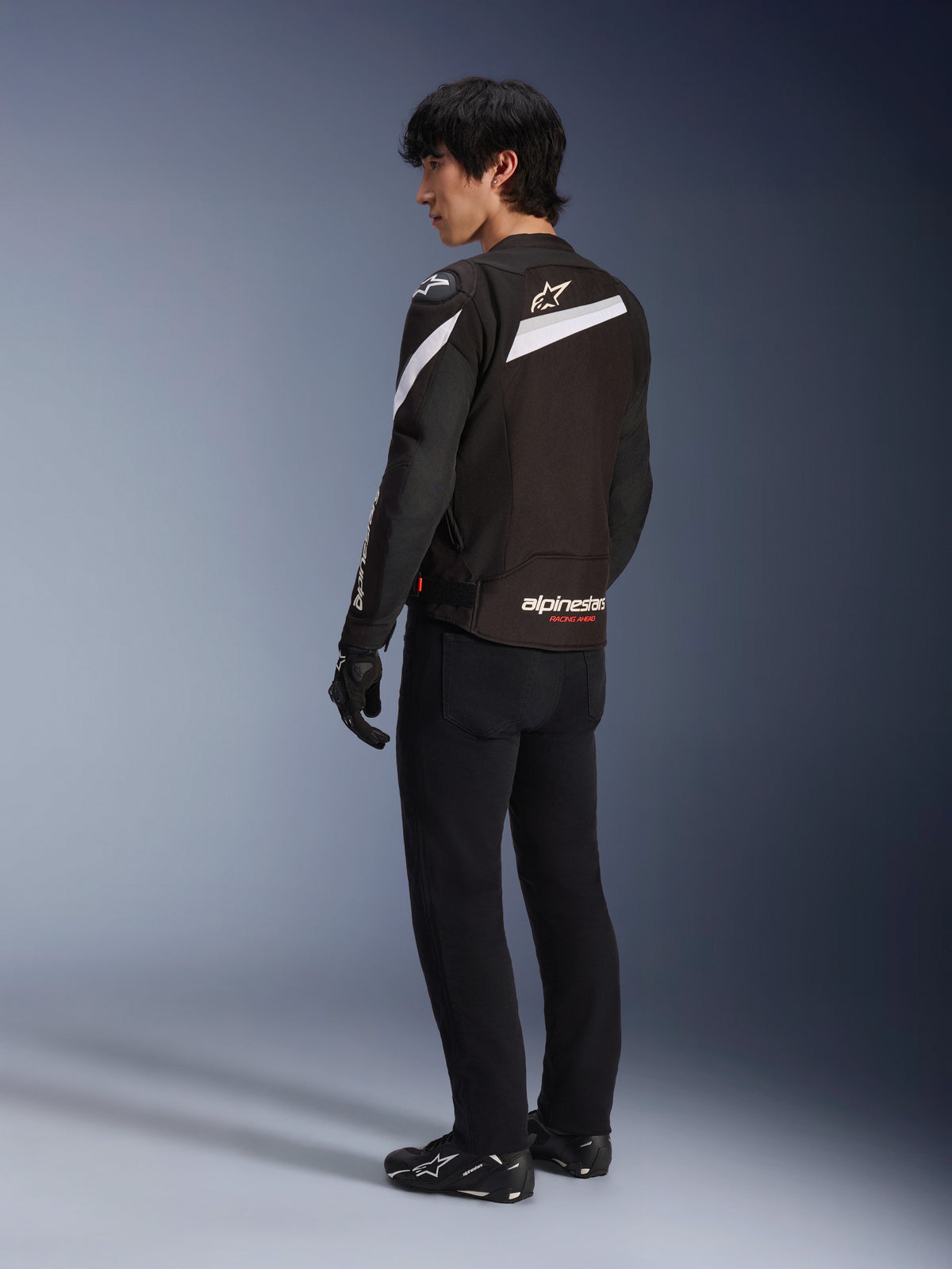 Piloto masculino vistiendo la chaqueta Alpinestars T-GP Plus R V4 en negro y blanco, vista trasera que muestra los acentos diagonales blancos y el logotipo en la parte inferior de la espalda, combinada con pantalones de conducción negros, guantes y botas técnicas, equipamiento textil de moto de ajuste deportivo