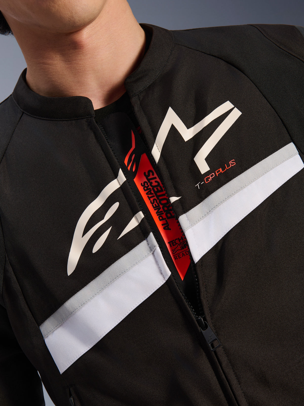 Chaqueta Alpinestars T-GP Plus R V4, primer plano del pecho y el cuello, tejido negro con logotipo de estrella blanca y marca T-GP PLUS, franja horizontal blanca y gris, cremallera abierta que revela una lengüeta interior roja con el texto Tech-Air Ready