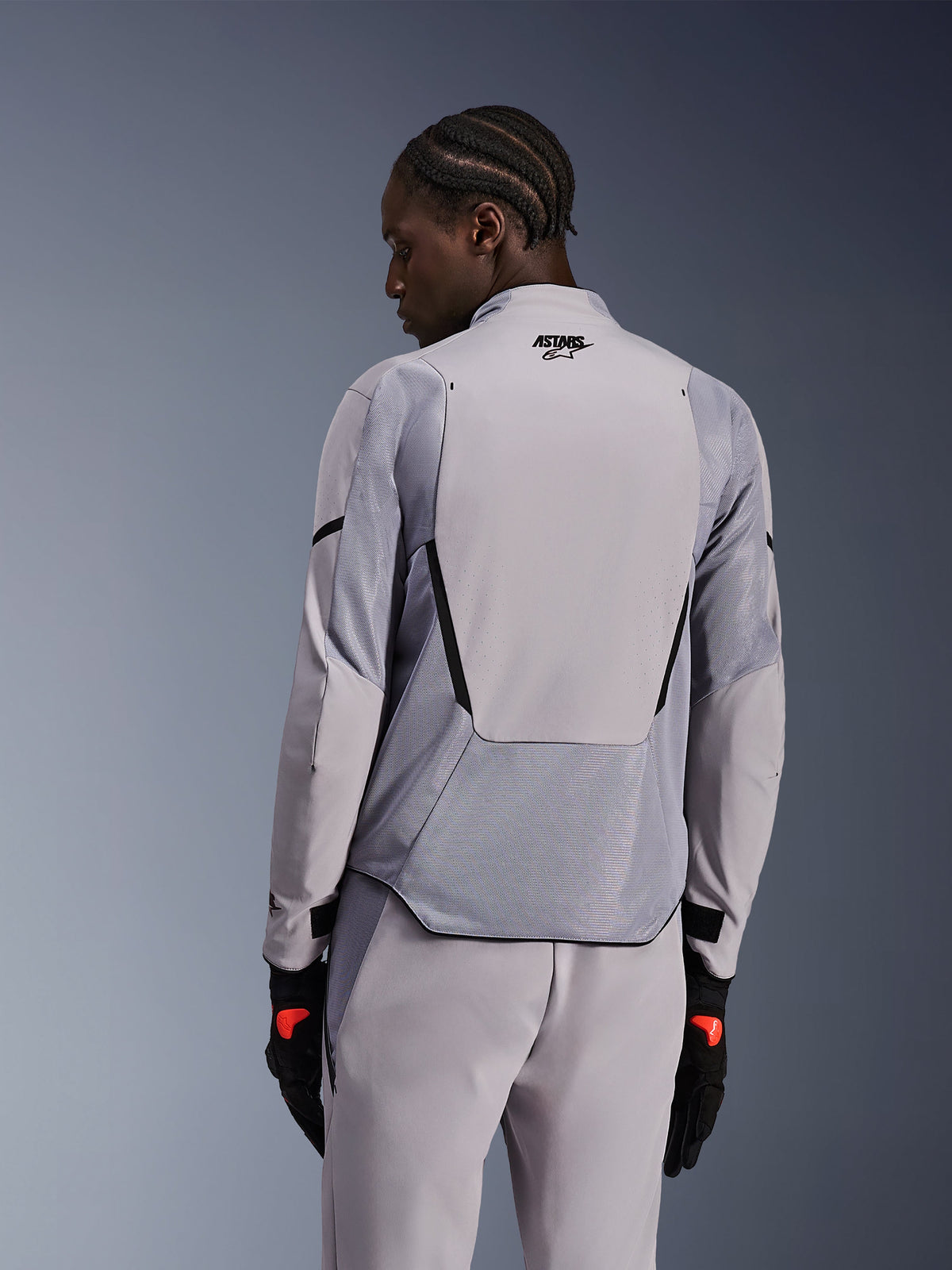 Chaqueta Alpinestars Aeroshell Airflow en Mid Gray Anthracite, vista trasera que muestra paneles de malla transpirable y perforaciones láser, construcción técnica de softshell elástico con un panel trasero alargado, combinada con pantalones grises a juego y guantes negros para un look deportivo.