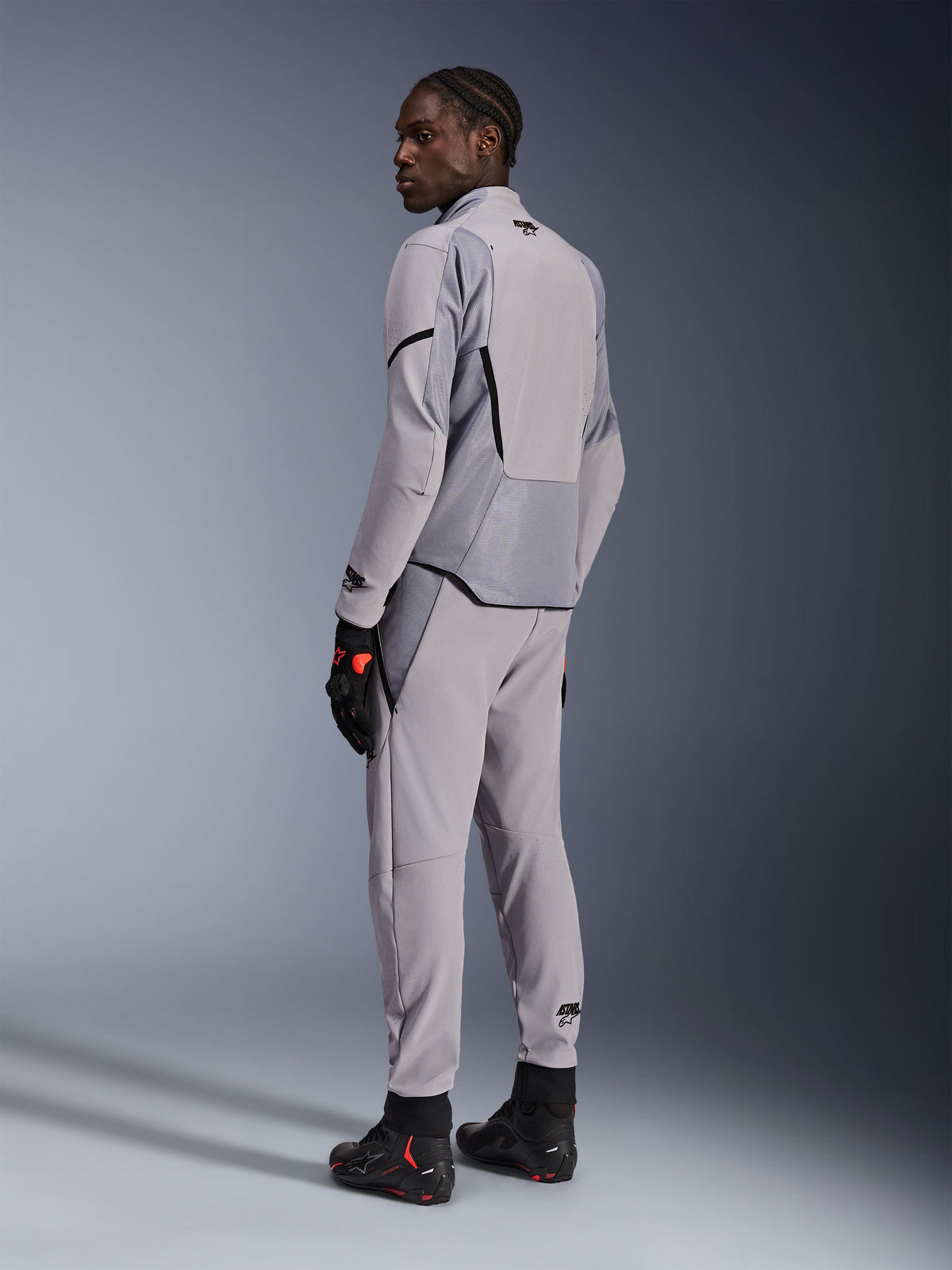 Chaqueta Alpinestars Aeroshell Airflow en color Mid Gray Anthracite, combinada con pantalones a juego y guantes de carreras negros, vista trasera que muestra los paneles perforados por láser y el ajuste deportivo, equipamiento profesional para motociclismo