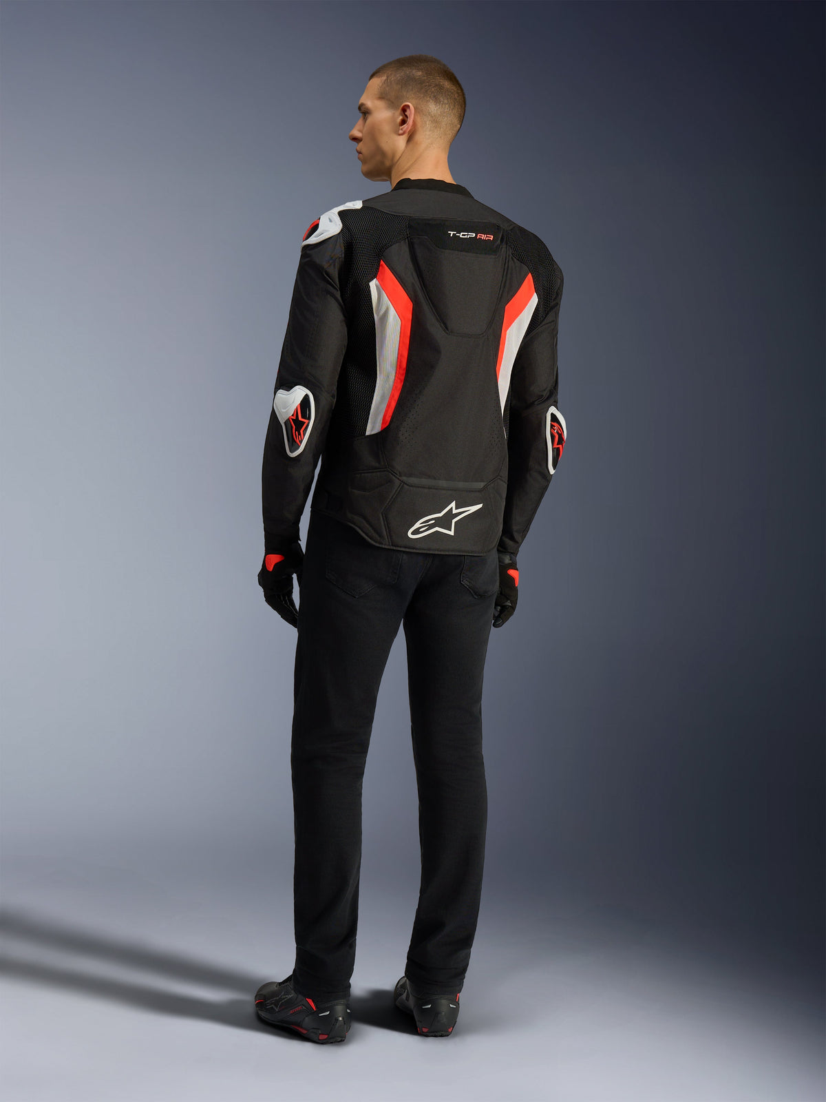 Alpinestars T-GP Air Jacket, chaqueta de moto en color Negro Rojo Fluo, vista trasera en un modelo, con paneles de ventilación de malla, joroba aerodinámica y protectores externos blancos en codos y hombros, combinada con pantalones negros y calzado de conducción