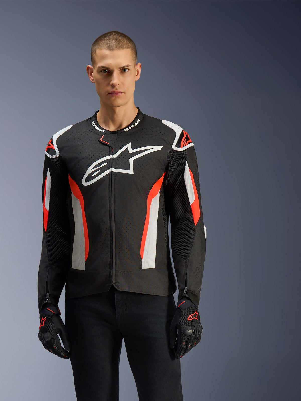 Modelo masculino con la chaqueta Alpinestars T-GP Air, negra con detalles en rojo flúor y blancos, logotipo de Alpinestars grande en el pecho, con paneles perforados por láser y protecciones en los hombros, combinada con guantes de moto negros, equipamiento de motociclismo de corte deportivo para climas cálidos