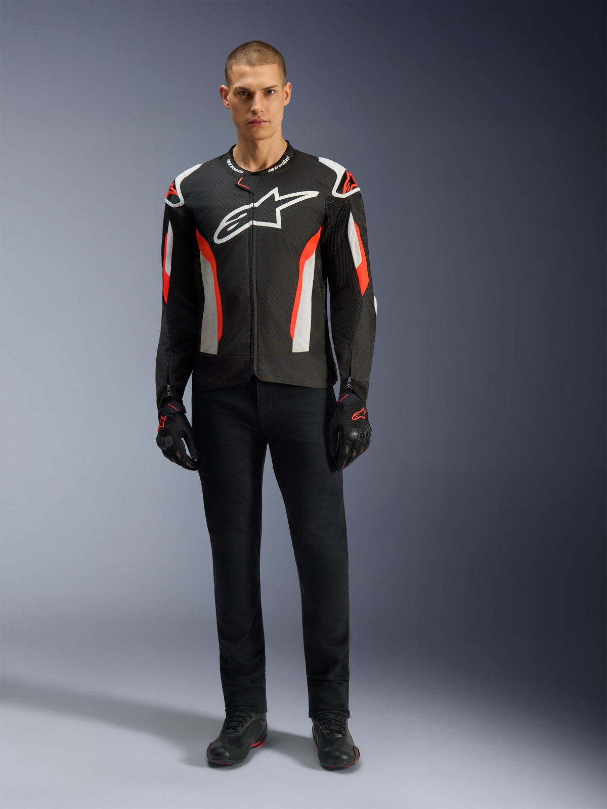Modelo masculino con la chaqueta Alpinestars T-GP Air en Black Red Fluo, chaqueta de motociclismo textil con detalles en blanco y rojo fluorescente, logotipo grande de Alpinestars en blanco en el pecho, paneles de ventilación de malla, combinada con pantalones técnicos negros y guantes de conducción, equipamiento con corte deportivo