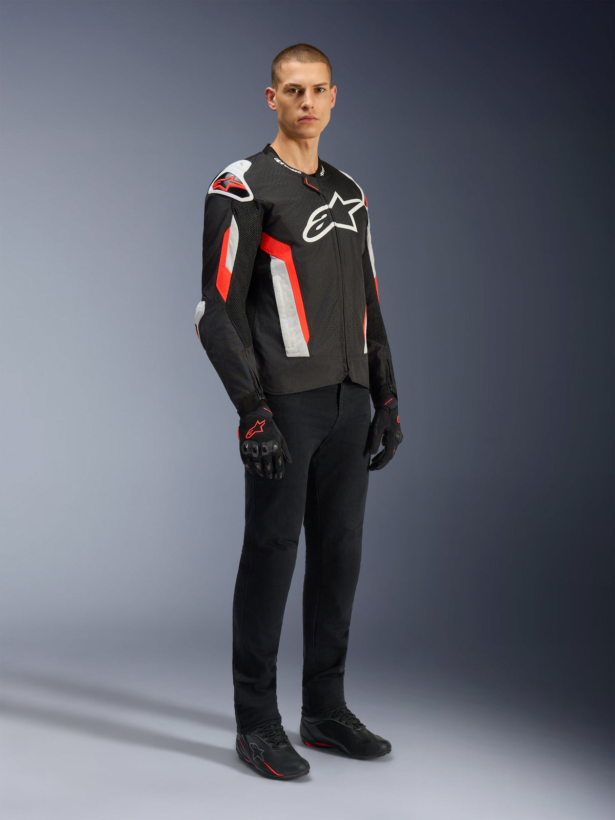 Chaqueta Alpinestars T-GP Air, Negro Rojo Flúor, usada por un modelo con pantalones de conducción y guantes negros, con paneles textiles perforados por láser, logotipos de Alpinestars en blanco y detalles en rojo, chaqueta de moto deportiva para carretera en climas cálidos