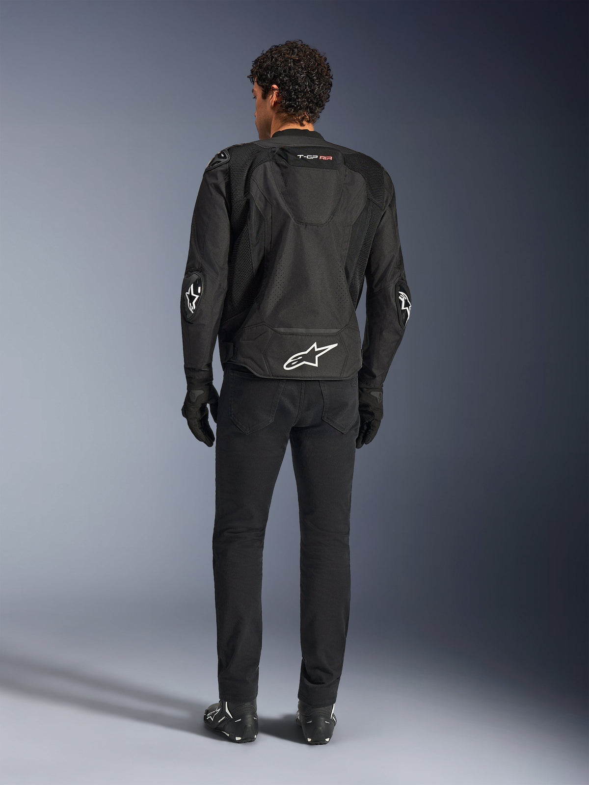 Alpinestars T-GP Air Jacket, Black, rear view on model, showing 3D mesh ventilation panels and aerodynamic hump, featuring white star logo on lower back and elbows, worn with black motorcycle pants and gloves, sport riding apparel.