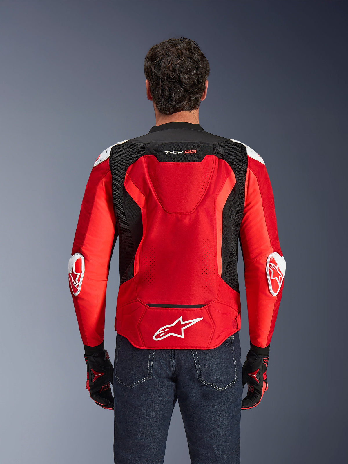 Chaqueta Alpinestars T-GP Air, chaqueta textil para moto, rojo brillante y negro con detalles en blanco, vista posterior que muestra la joroba aerodinámica y paneles de ventilación de malla, con el logotipo de la estrella de Alpinestars y la marca T-GP Air, usada con vaqueros oscuros y guantes de carreras.
