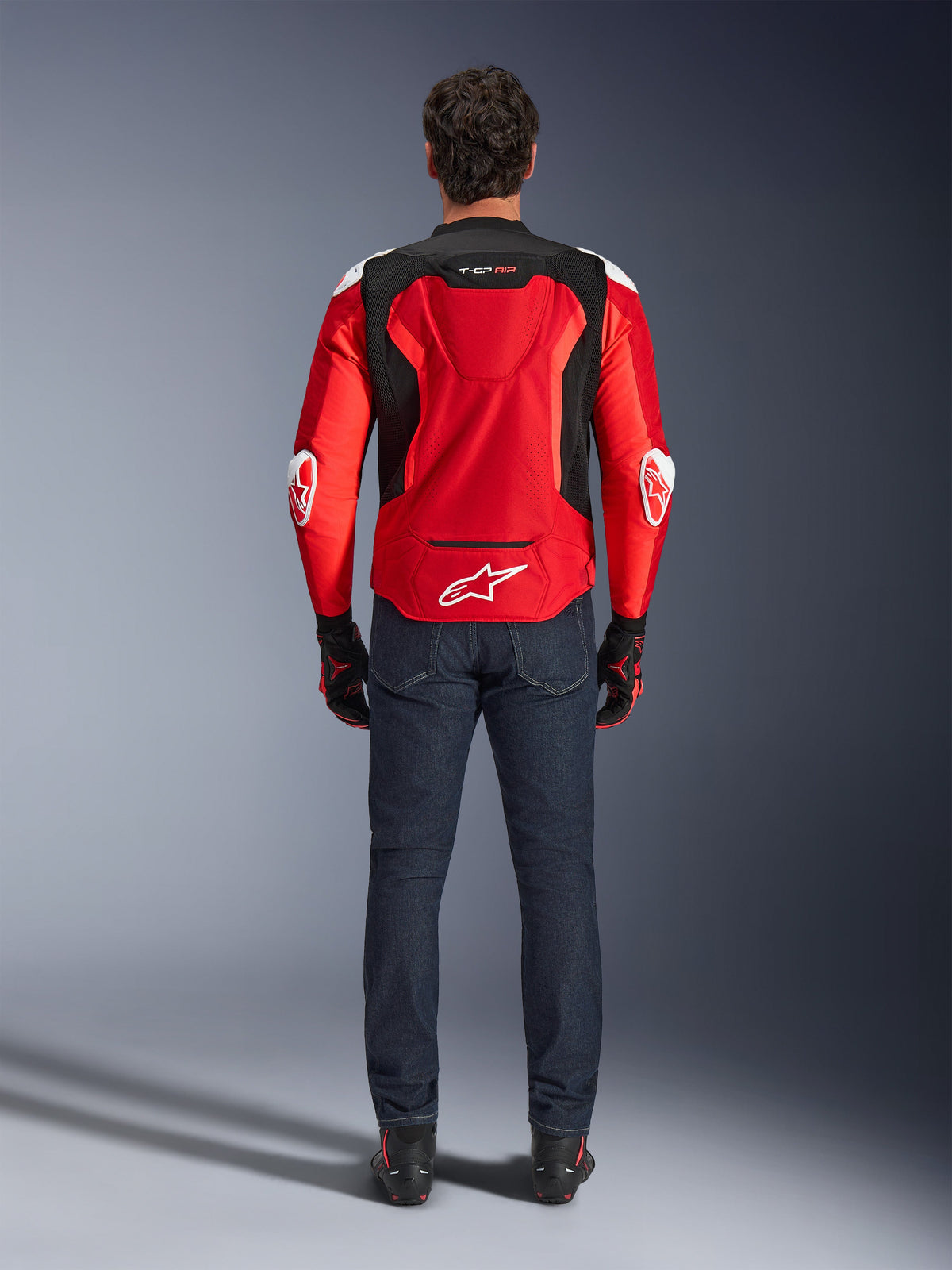 Chaqueta Alpinestars T-GP Air, vista trasera, rojo brillante y negro, con logotipo blanco en la parte baja de la espalda y marca 'T-GP AIR', usada con vaqueros de color azul oscuro y guantes de moto negros y rojos, equipamiento de motociclismo de alto rendimiento deportivo