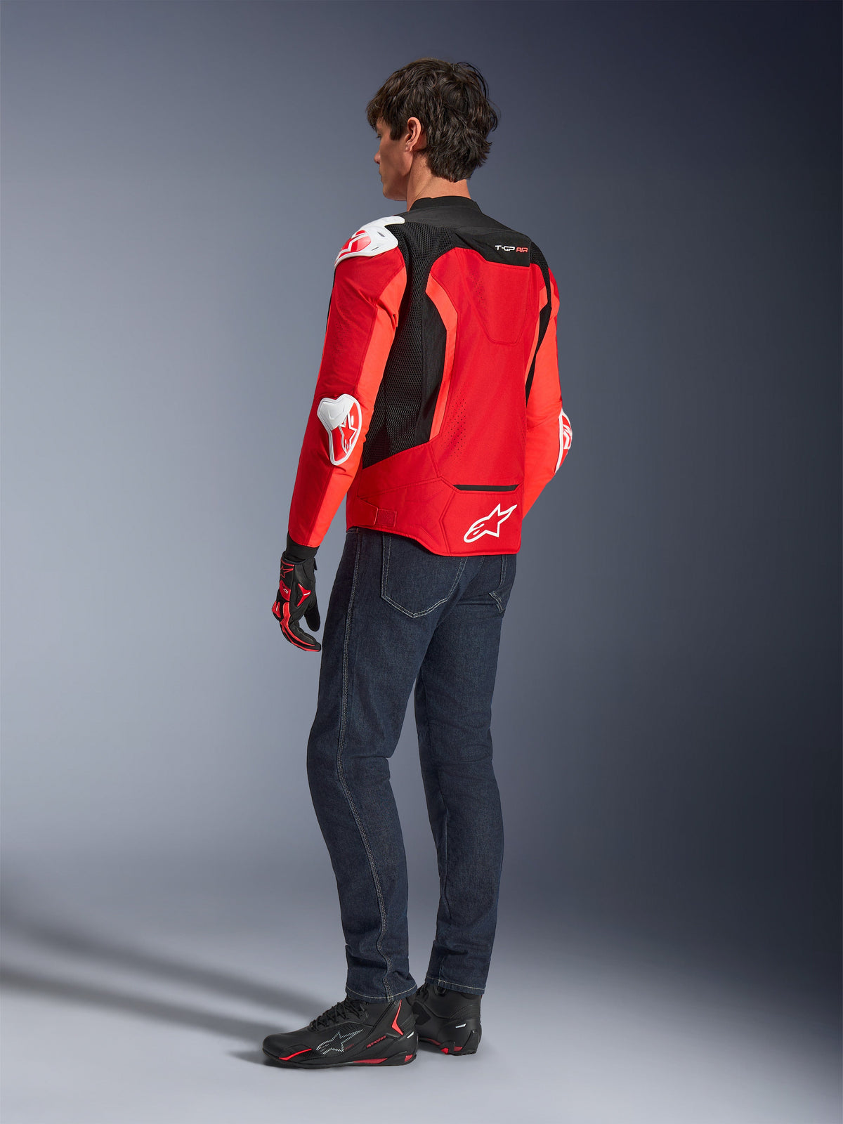 Chaqueta Alpinestars T-GP Air, vista trasera de un hombre con una chaqueta textil en rojo brillante y rojo flúor con paneles de malla negros y deslizadores blancos, combinada con vaqueros azul oscuro y calzado de moto negro, equipamiento deportivo para motociclismo.