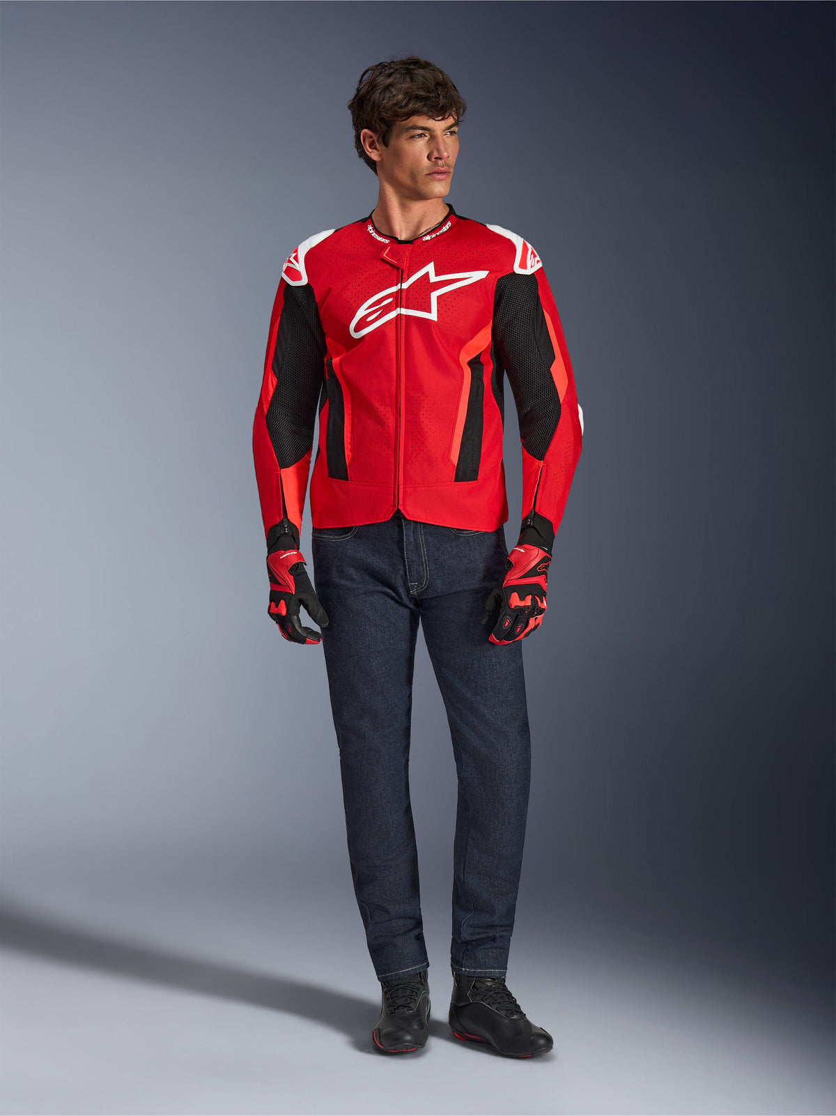 Chaqueta Alpinestars T-GP Air, rojo brillante, rojo flúor y negro, modelo masculino con equipamiento de moto completo, vaqueros azul oscuro y guantes a juego, chaqueta textil deportiva con paneles de malla perforada y logo Astars blanco, foto de estudio.