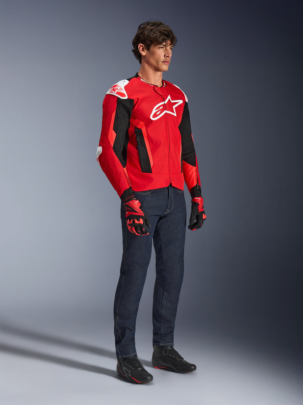 Chaqueta Alpinestars T-GP Air, chaqueta deportiva para moto, rojo brillante y negro con detalles en blanco, usada por un modelo con jeans azul oscuro y guantes de carreras rojos, con paneles de malla de alto flujo y deslizadores de hombro blancos, ajuste deportivo aerodinámico para conducir en climas cálidos