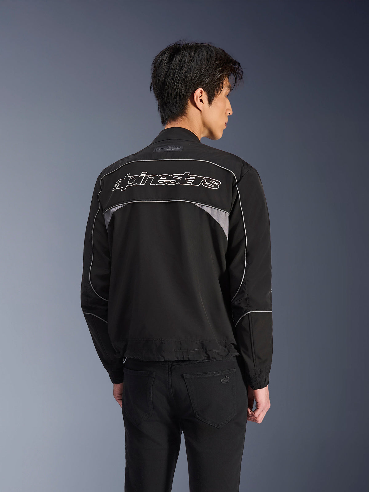 Chaqueta Alpinestars Unite, chaqueta de moto urbana, negra con detalles en gris oscuro, vista trasera que muestra el logotipo de Alpinestars en blanco sobre los hombros, ribetes y paneles reflectantes, diseño softshell minimalista estilo streetwear