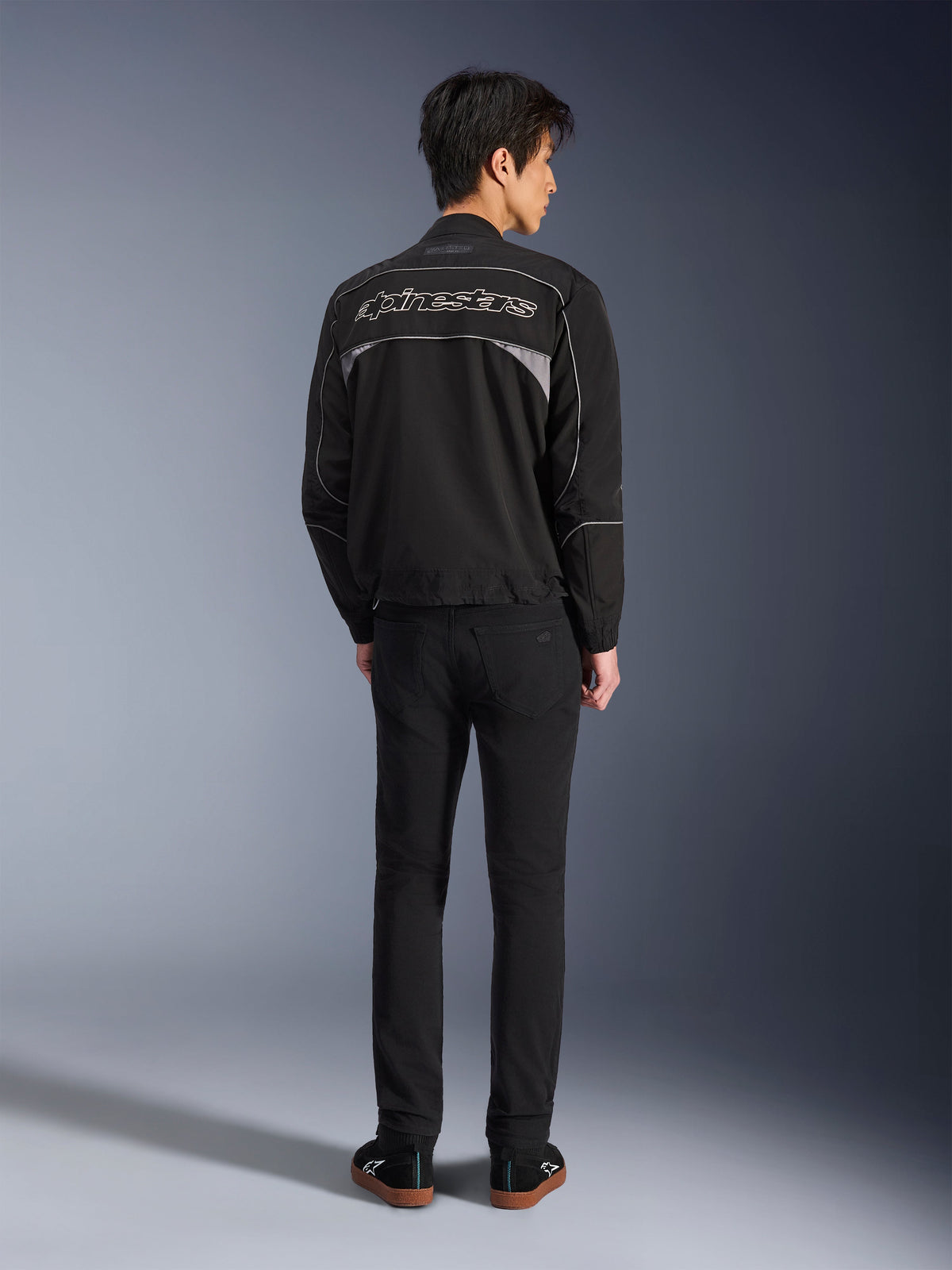 Chaqueta Alpinestars Unite, chaqueta de moto negra y gris oscuro, vista trasera que muestra el logotipo de Alpinestars perfilado en la espalda, paneles reflectantes grises, diseño urbano minimalista, combinada con pantalones negros y calzado negro.