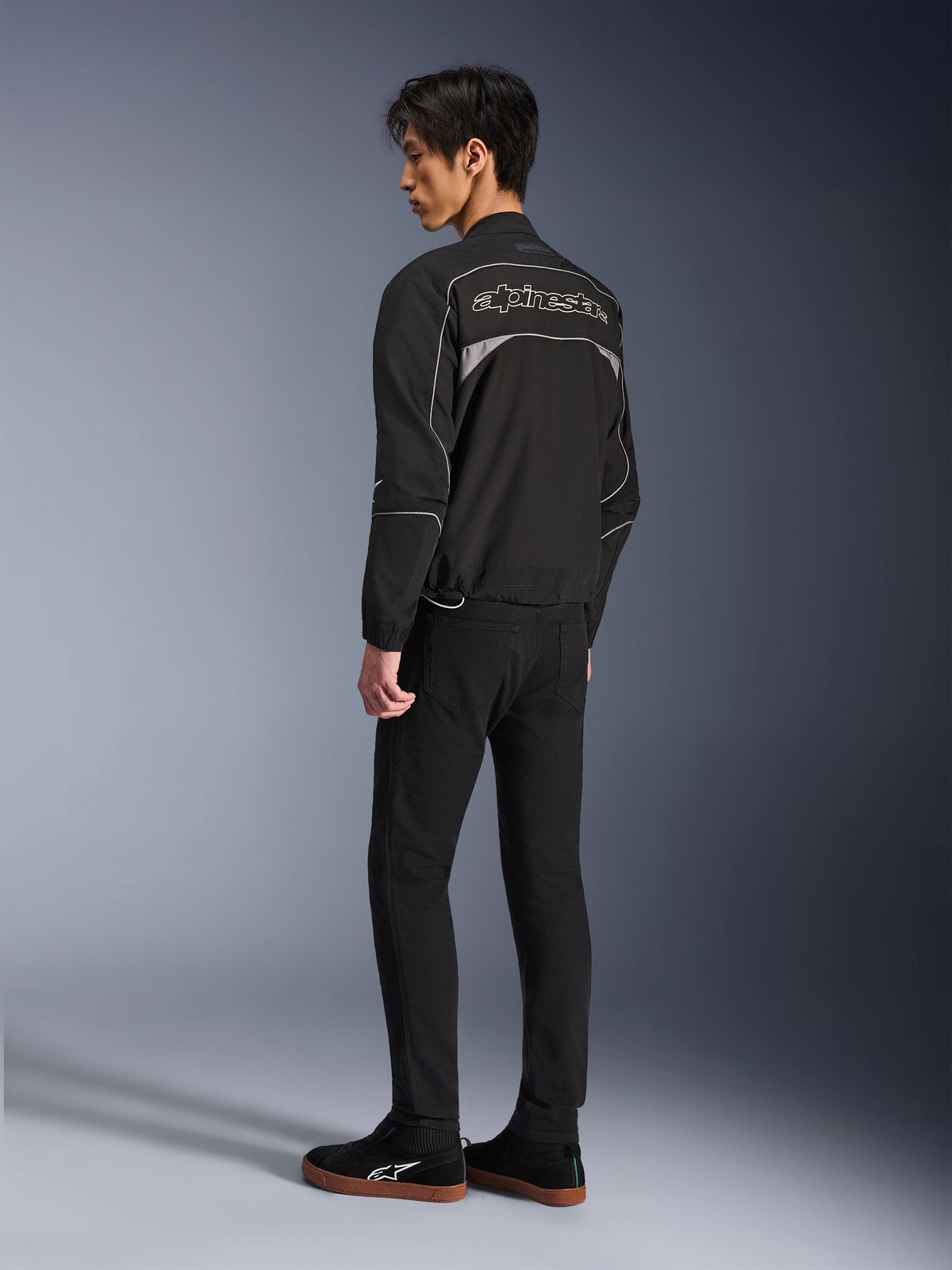Chaqueta Alpinestars Unite, chaqueta de moto urbana, negro y gris oscuro, vista trasera que muestra un gran logotipo de Alpinestars perfilado, ribetes y paneles reflectantes, diseño softshell minimalista, usada con pantalones negros y calzado casual