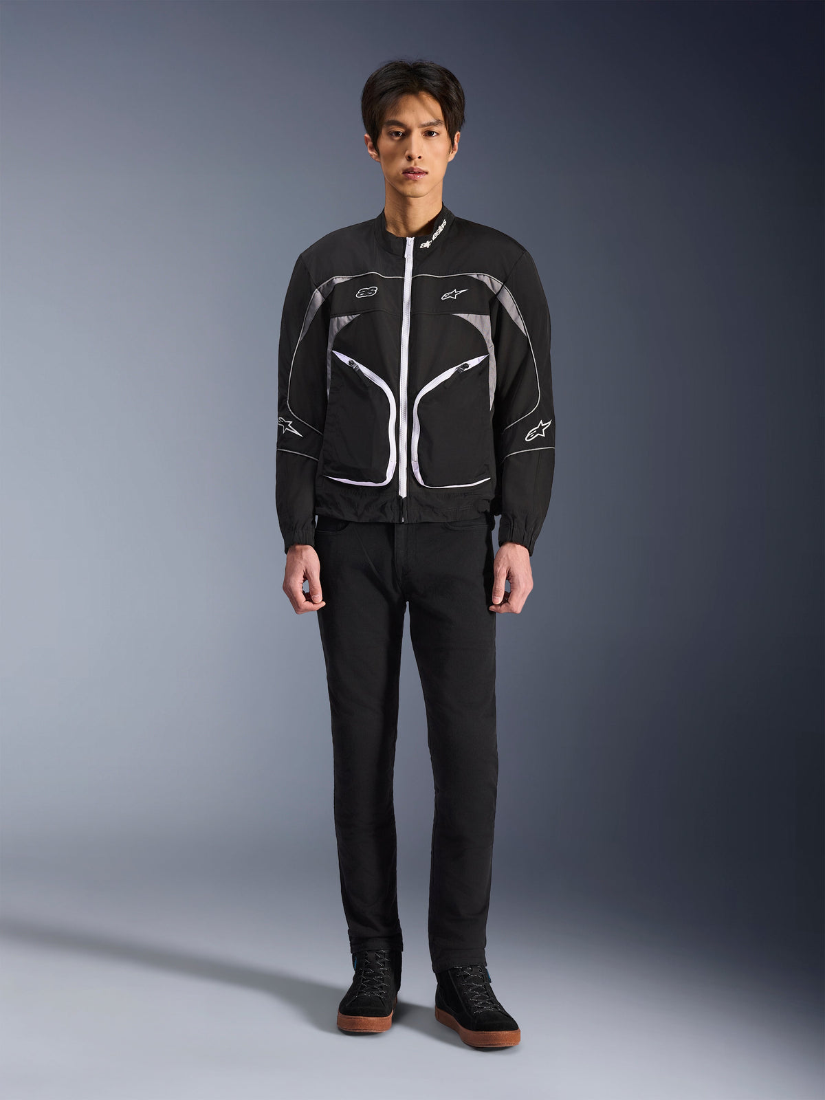 Chaqueta Alpinestars Unite, chaqueta de moto, negro y gris oscuro, usada por un modelo con pantalones negros y botas negras, con cremalleras blancas en contraste, ribetes reflectantes y diseño urbano minimalista