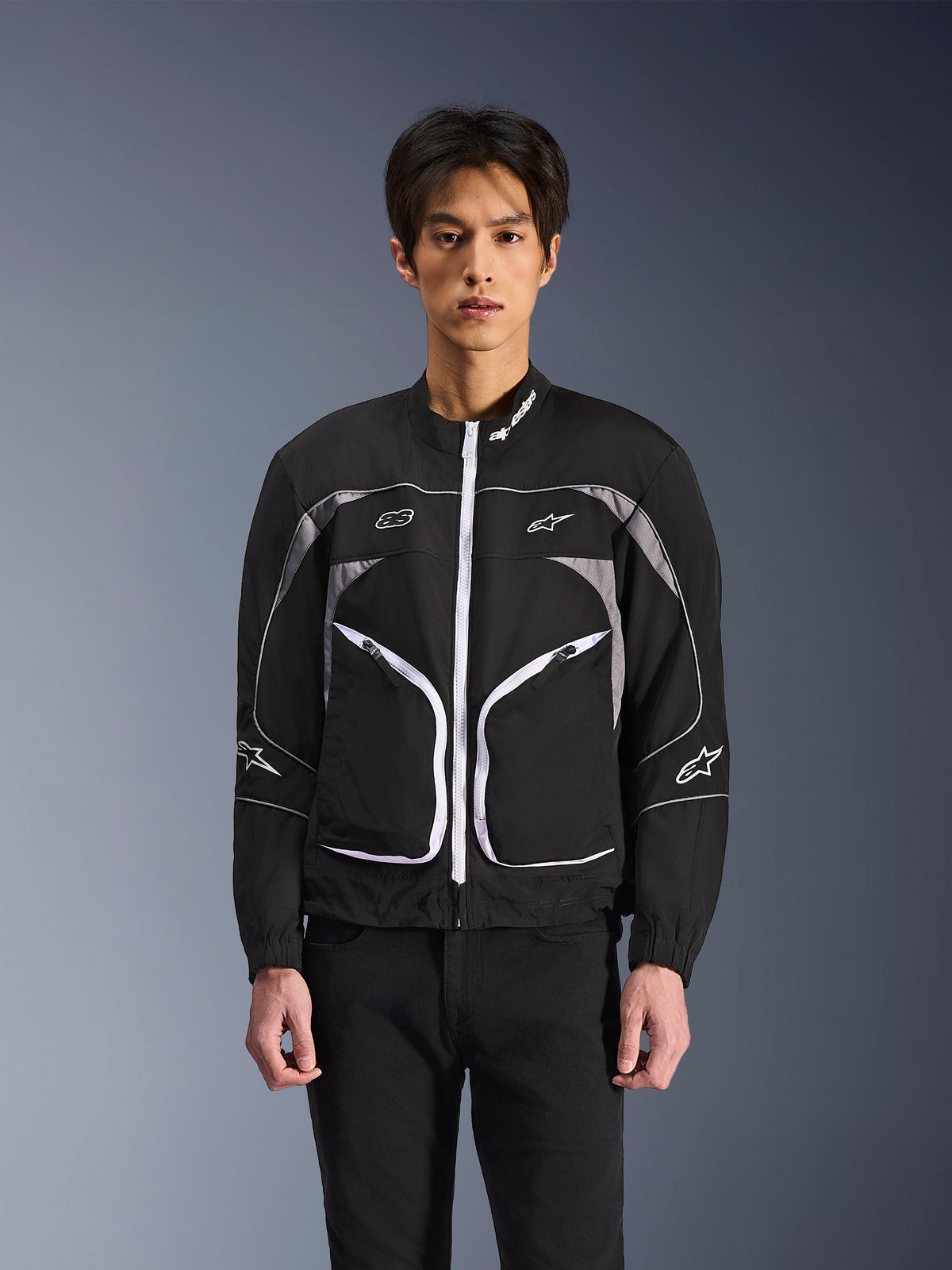 Chaqueta Alpinestars Unite, chaqueta de moto urbana y minimalista, negro y gris oscuro con detalles blancos, presenta una cremallera frontal blanca, ribetes reflectantes y logotipos de Alpinestars en el pecho y las mangas, estilo streetwear contemporáneo para motoristas.