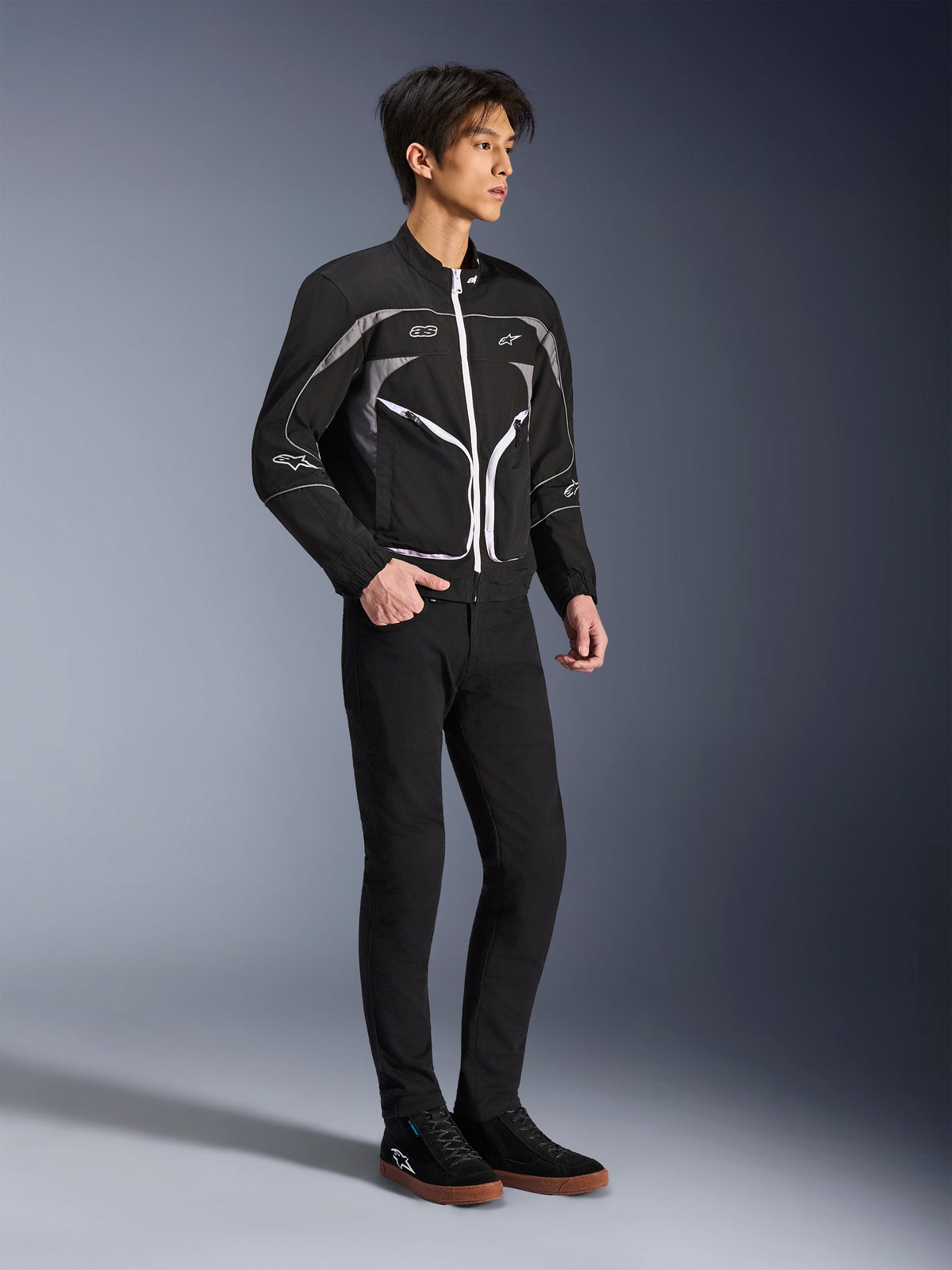 Modelo vistiendo la chaqueta Alpinestars Unite en negro y gris oscuro, chaqueta urbana para motociclista con cremalleras blancas y detalles reflectantes, combinada con pantalones negros de corte ajustado y zapatillas, equipamiento de conducción minimalista de inspiración urbana.