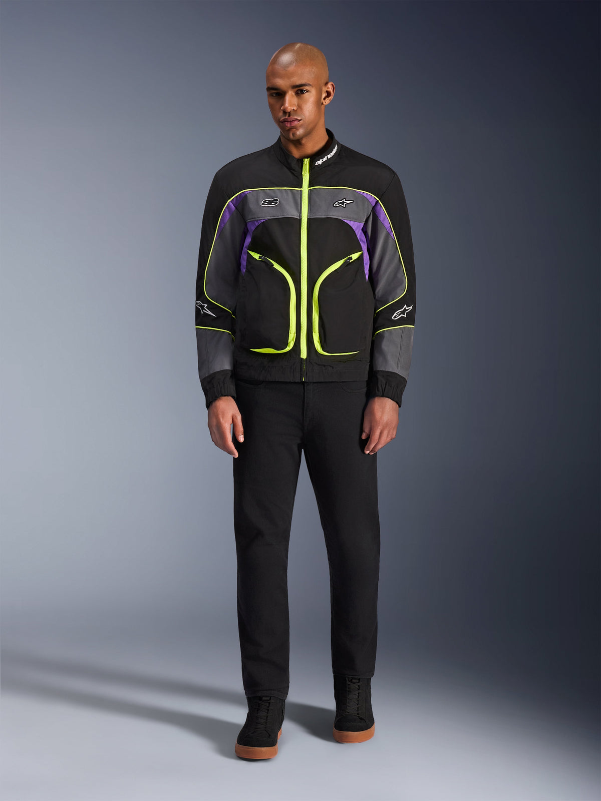 Male model wearing the Alpinestars Unite Jacket in Black Asphalt Violet, featuring a black base with grey and purple panels and vibrant neon yellow zippers and trim, paired with black pants and shoes, minimalist urban motorcycle jacket.