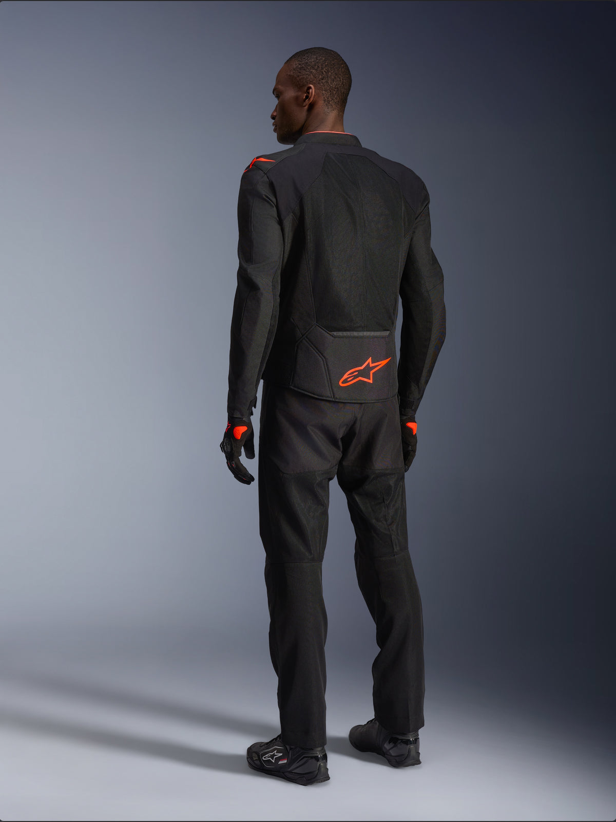 Male rider from the back wearing the Alpinestars Viper Air V4 Jacket, Black and Red Fluo, featuring an orange Astars logo on the lower back and shoulders, mesh textile construction, styled with matching black motorcycle pants and boots.