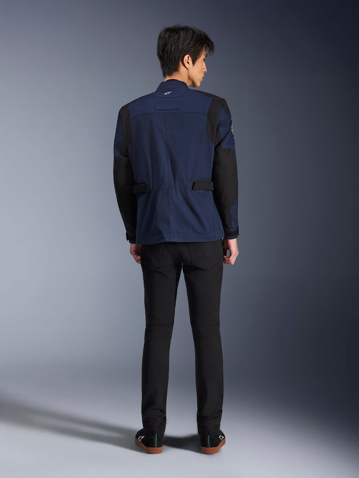 Modelo vistiendo la chaqueta Alpinestars Dice en azul oscuro y negro, vista posterior, chaqueta de motociclismo urbana tipo field jacket con hombros de pliegue estructural y pestañas de ajuste en la cintura, combinada con pantalones negros y zapatillas negras.