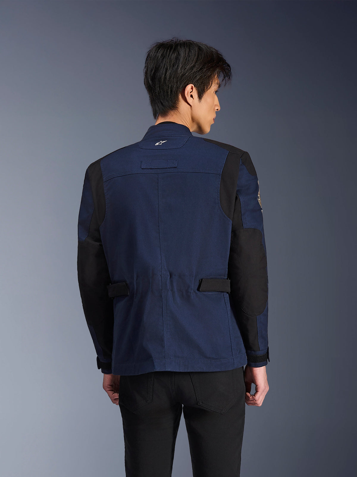 Alpinestars Dice Jacket, chaqueta de moto urbana estilo field en azul oscuro y negro, vista posterior usada por un hombre, con paneles negros reforzados en hombros y codos y tiras de ajuste en la cintura.