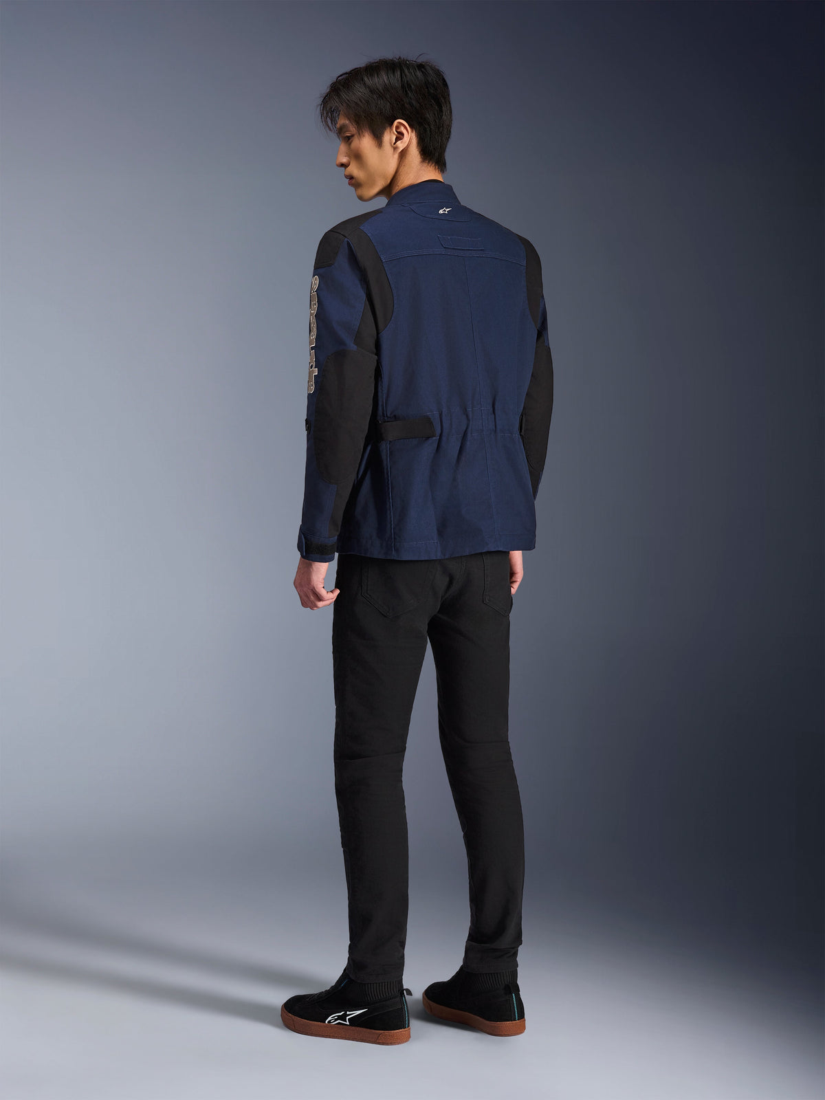 Modelo masculino visto desde atrás vistiendo la Chaqueta Dice de Alpinestars en azul oscuro y negro, combinada con pantalones negros y zapatillas negras; chaqueta de moto urbana tipo field jacket con hombros de pliegues estructurales y ajustadores de cintura.
