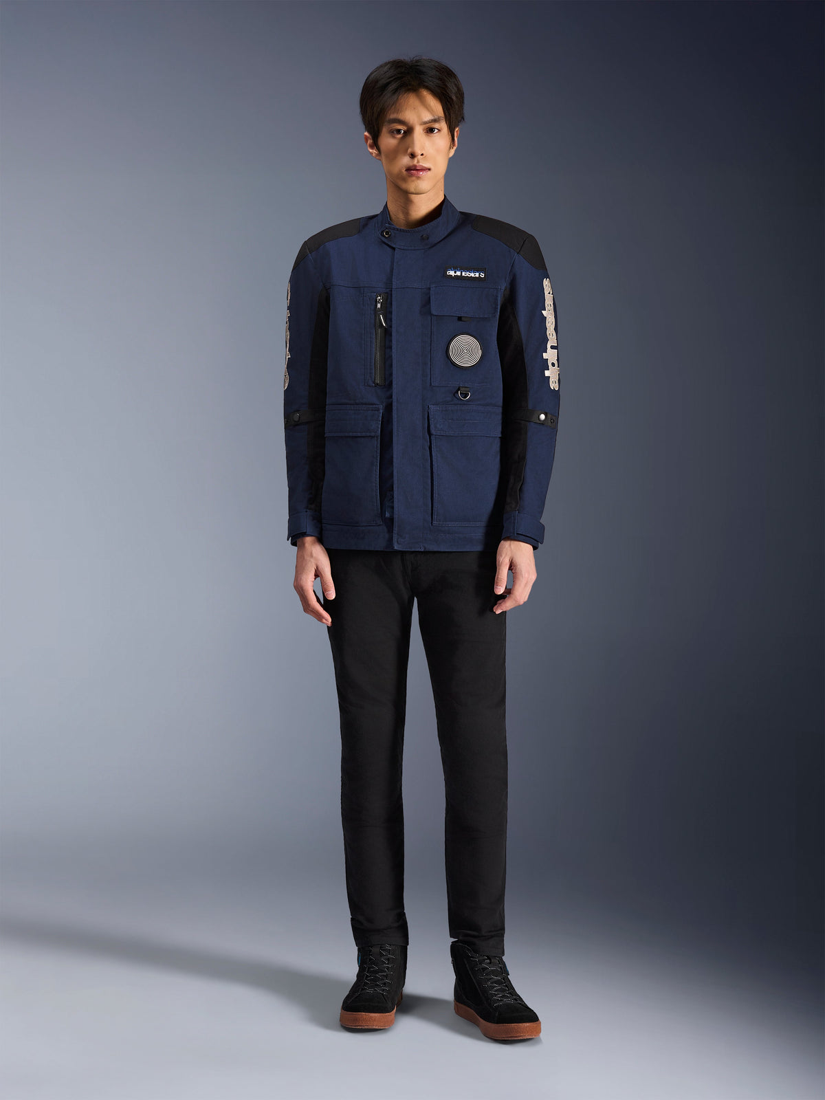 Chaqueta Alpinestars Dice, chaqueta de moto urbana, azul oscuro y negro, usada por un modelo con pantalones negros y calzado negro, diseño moderno de chaqueta de campo con múltiples bolsillos, cremallera vertical en el pecho y logotipo de Alpinestars en blanco en las mangas