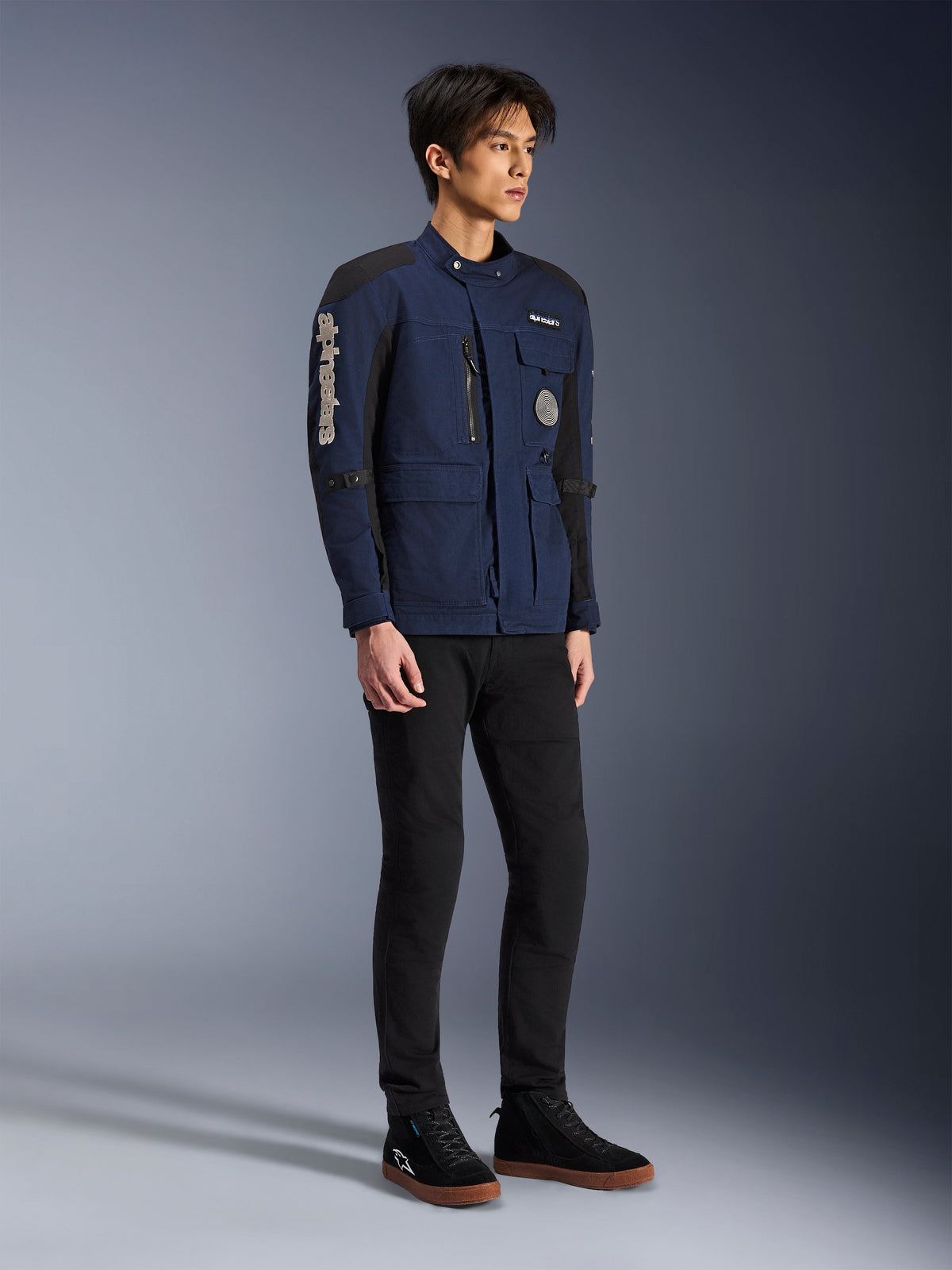 Chaqueta Alpinestars Dice, azul oscuro y negro, chaqueta de moto moderna estilo field usada por un modelo con pantalones negros y zapatillas altas negras, con múltiples bolsillos de utilidad, marca Alpinestars vertical en la manga y parches tejidos, equipo de conducción urbano contemporáneo