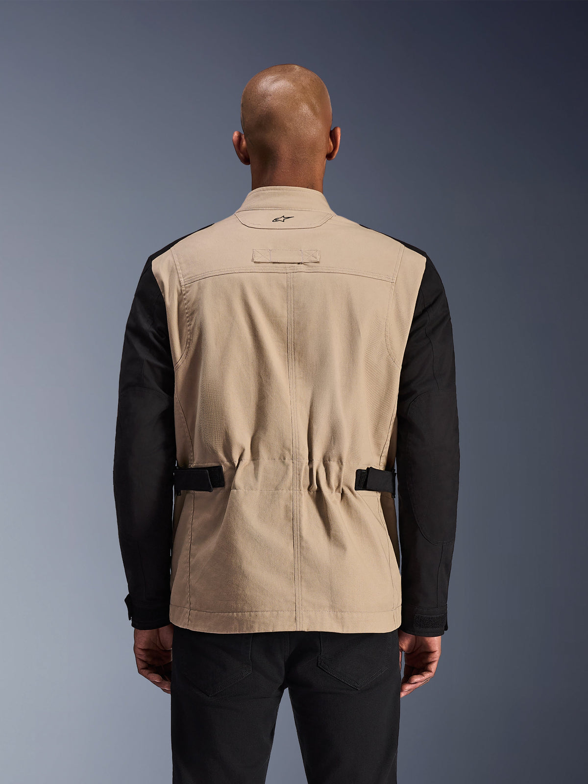 Chaqueta Alpinestars Dice, chaqueta de moto urbana, aluminio y negro, vista trasera en un modelo, con hombros plisados para movilidad, correas de cintura ajustables y construcción robusta de sarga de policotón.