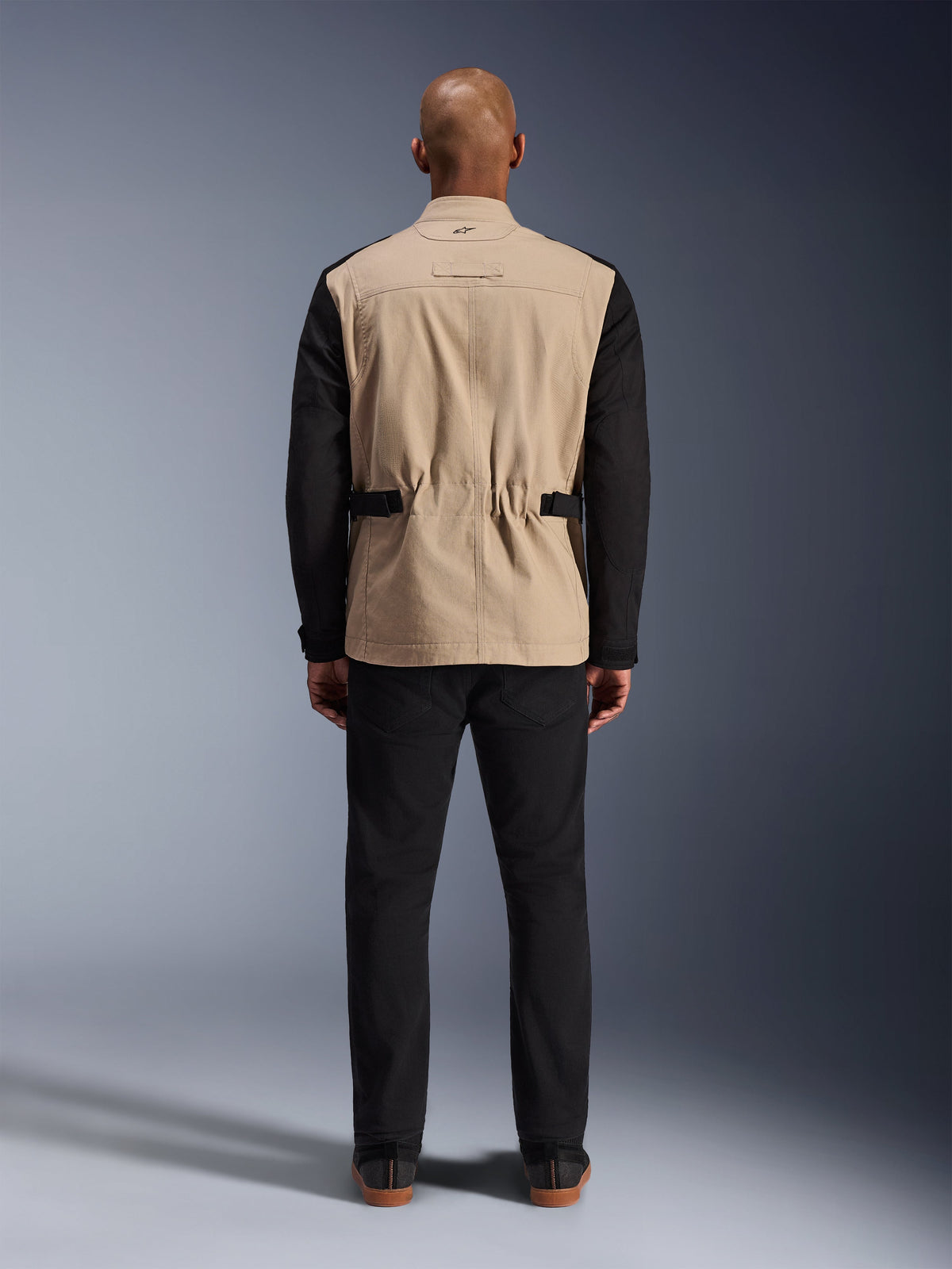 Chaqueta Dice de Alpinestars, chaqueta urbana para moto, beige y negro, vista posterior en modelo, con confección en sarga de policotón, correas de ajuste en la cintura y un pequeño logotipo bordado en el cuello, combinada con pantalones negros