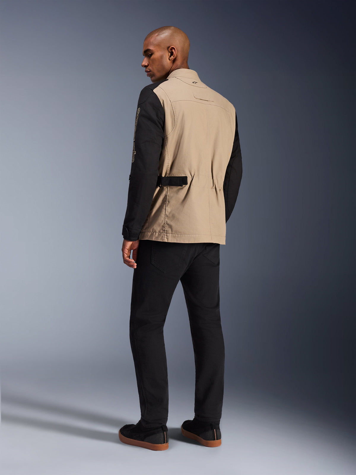 Alpinestars Dice Jacket, chaqueta de moto urbana, color Aluminum Black con torso canela y mangas negras, vista trasera en modelo, diseño robusto inspirado en ropa de trabajo con ajustadores de cintura, combinada con pantalones de conducción negros.