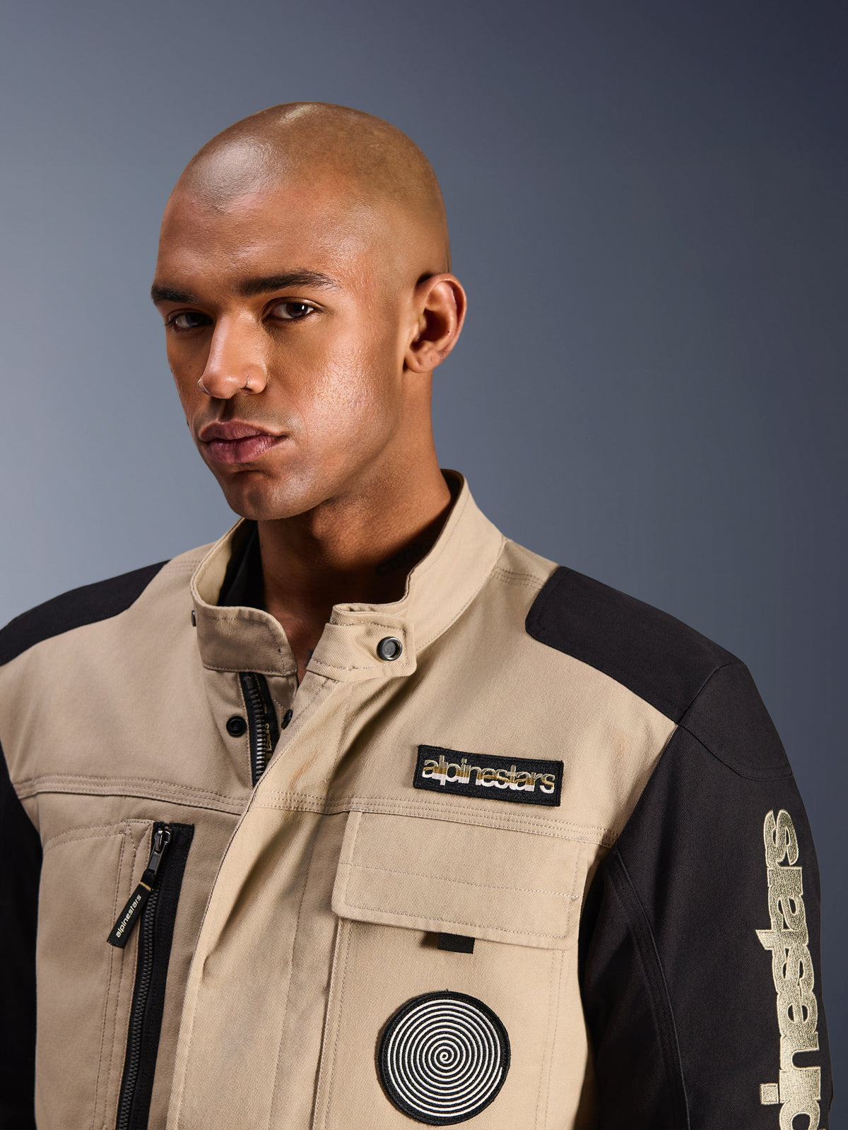 Modelo masculino con la chaqueta Alpinestars Dice en Aluminum Black, chaqueta urbana para moto con paneles de bloques de color canela y negro, con un parche bordado de Alpinestars en el pecho y logotipo en espiral, diseño robusto inspirado en la ropa de trabajo con cuello alto.