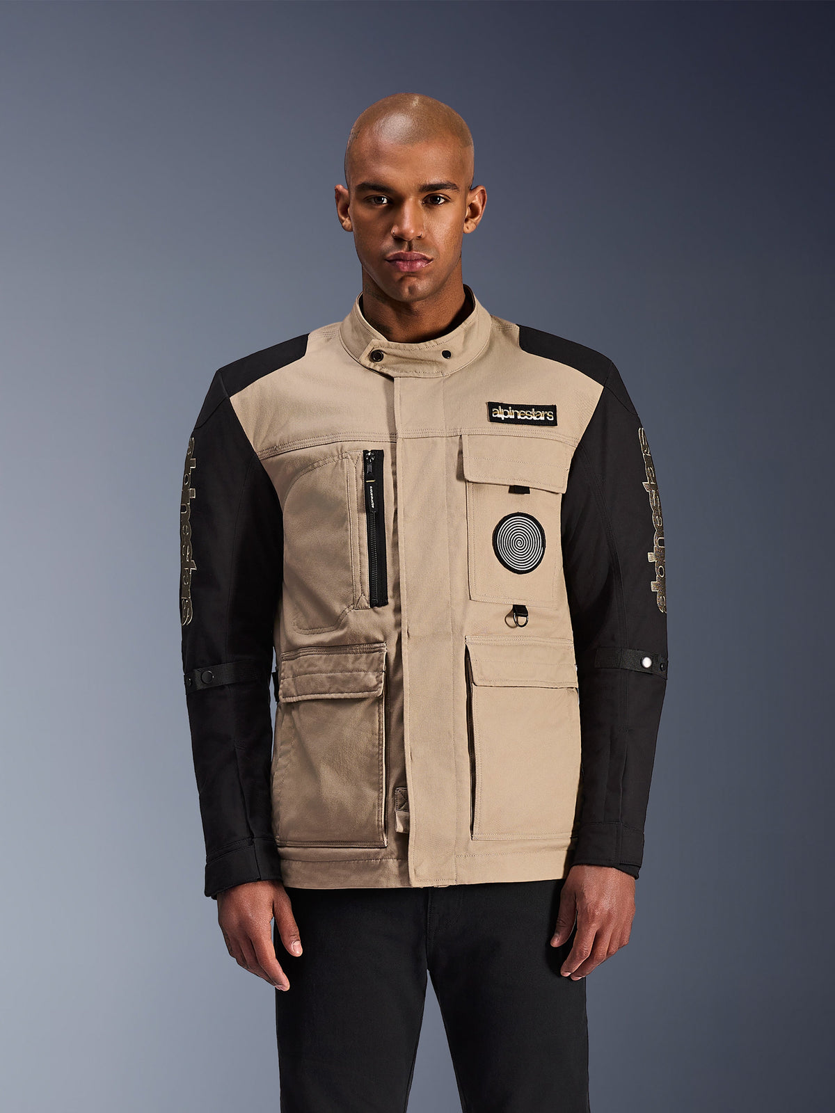 Alpinestars Dice Jacket, chaqueta de moto urbana, beige y negra, estética robusta de ropa de trabajo con múltiples bolsillos utilitarios y parches en el pecho, cuello alto con cierre de botón, logotipos llamativos en las mangas, usada por un modelo masculino