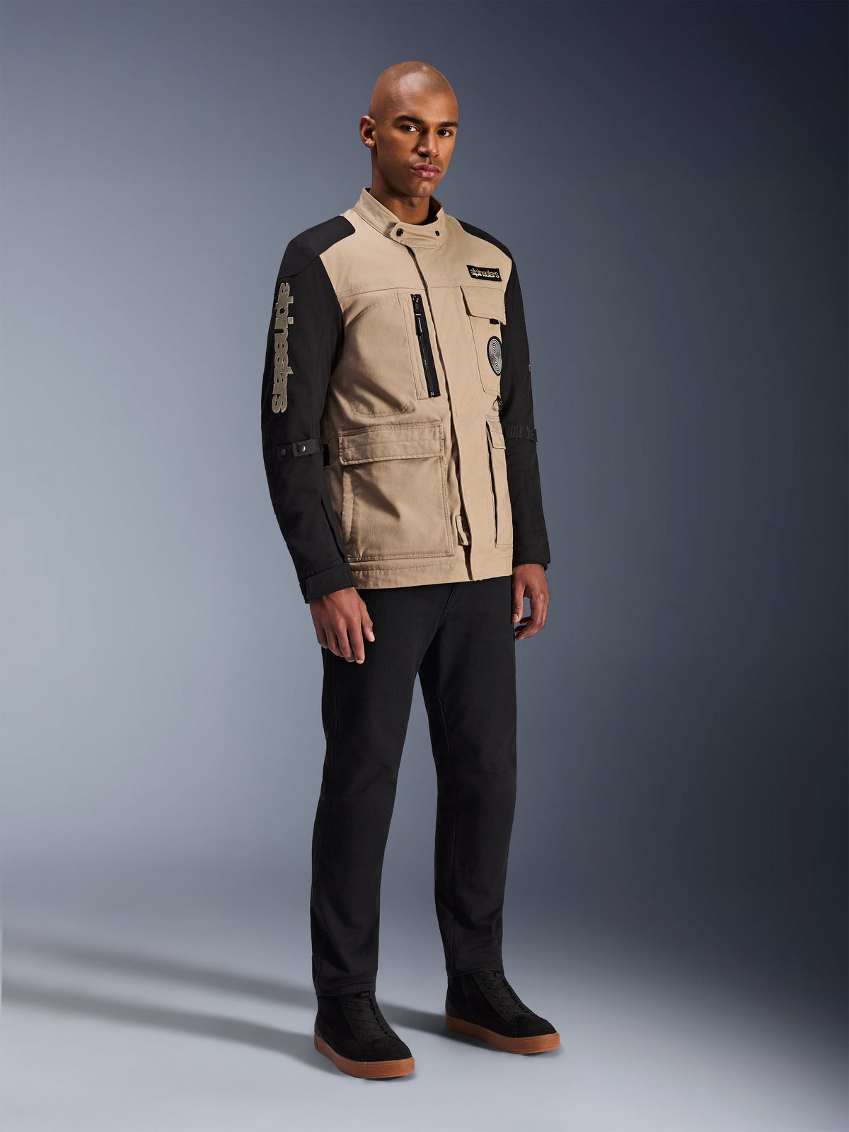 Chaqueta Alpinestars Dice, chaqueta urbana de motociclismo, Aluminum Black y tostado, diseño en dos tonos con mangas negras y torso caqui, lucida por un modelo con pantalones negros y zapatillas negras, estética de inspiración 'workwear' con bolsillos funcionales y marca bordada en la manga.