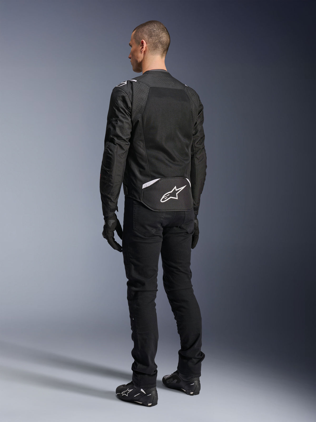 Chaqueta Alpinestars T-Sps Air V2, chaqueta de moto textil, negra, vista trasera que muestra un gran panel de ventilación de malla y el logotipo blanco de Alpinestars en la parte inferior de la espalda, combinada con pantalones y guantes negros para un estilo urbano deportivo.