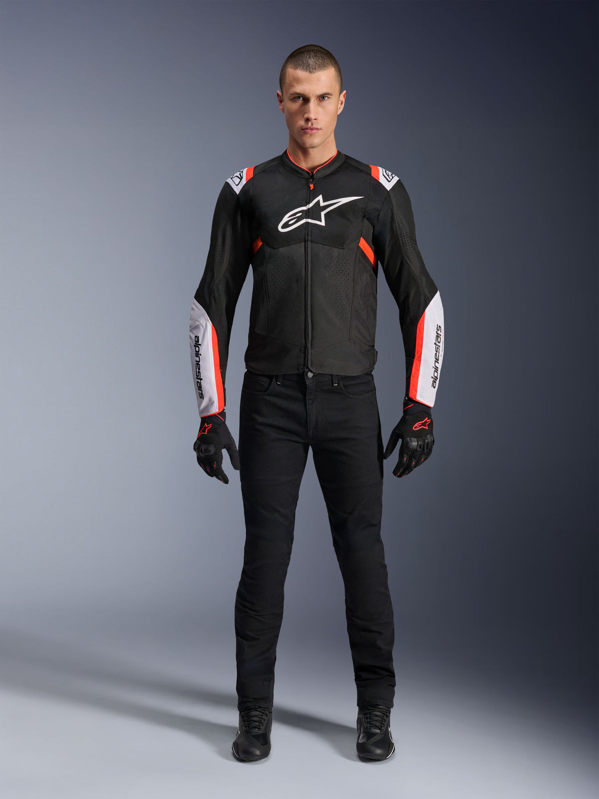 Modelo masculino con la chaqueta Alpinestars T-Sps Air V2 en negro, blanco y rojo flúor, con grandes paneles de malla técnica y logotipos blancos llamativos en el pecho y las mangas, combinada con pantalones técnicos de conducción y guantes negros, equipamiento deportivo urbano para moto