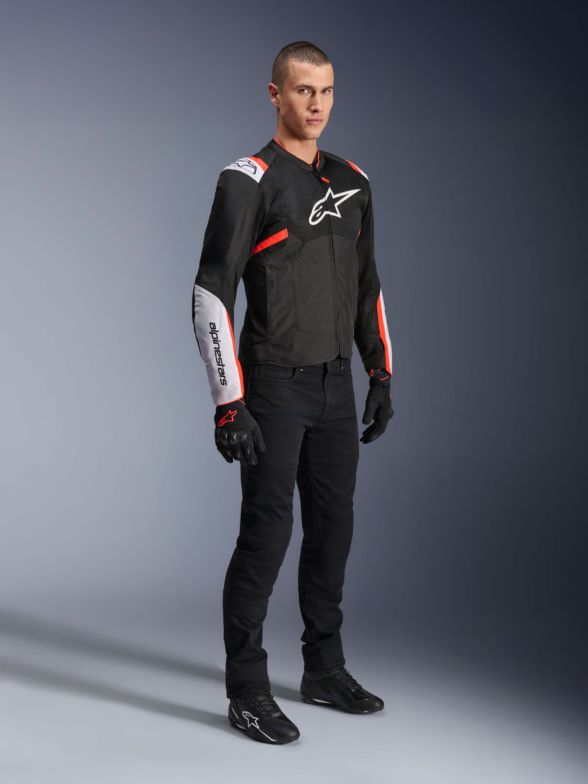 Chaqueta Alpinestars T-Sps Air V2, negro, blanco y rojo flúor, vestida por un modelo masculino con pantalones y guantes de moto negros, chaqueta textil de moto con amplios paneles de malla, secciones blancas en hombros y antebrazos, y un logotipo de Alpinestars destacado en el pecho.