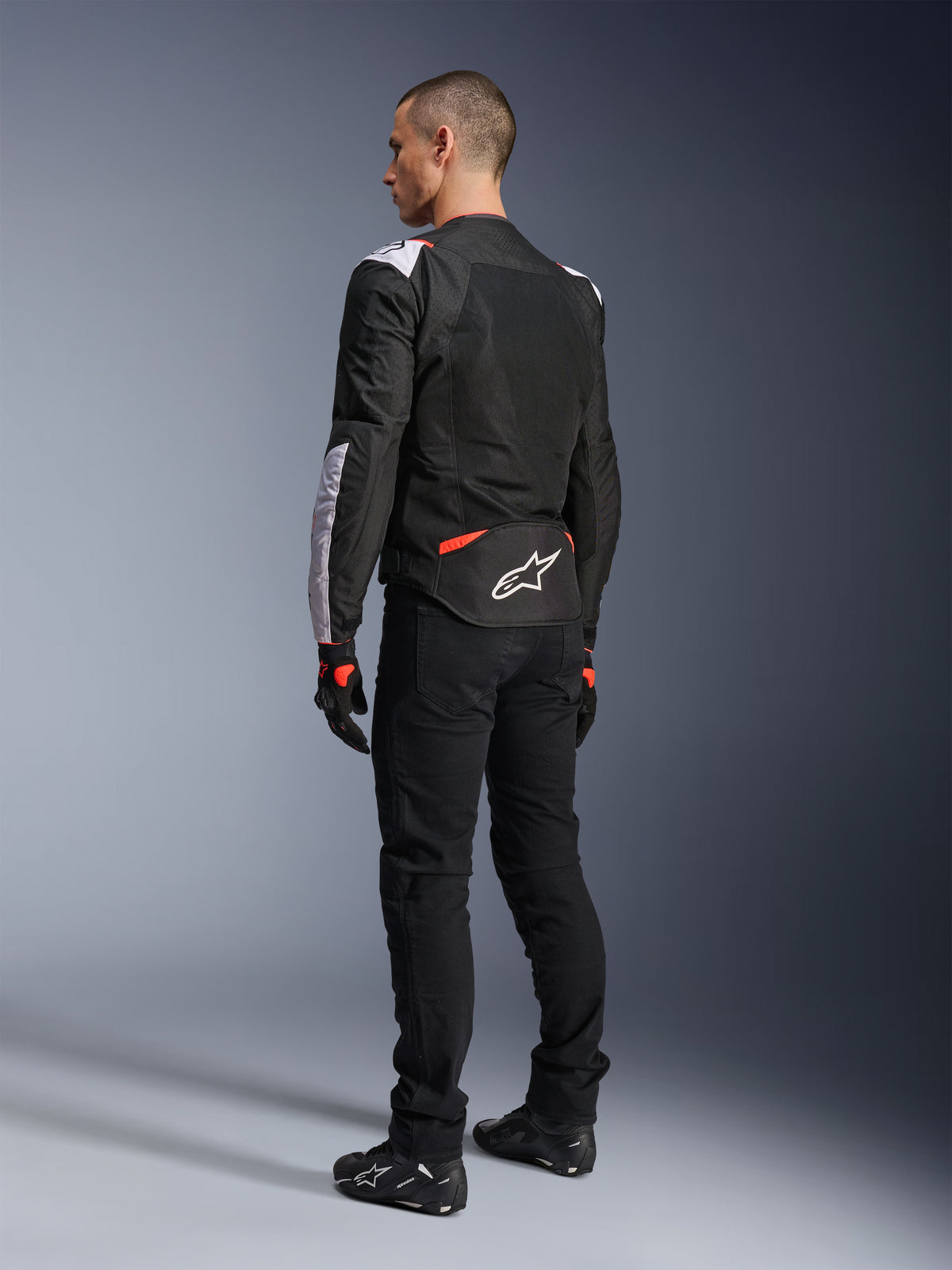 Chaqueta Alpinestars T-Sps Air V2, chaqueta textil de moto, negra, blanca y roja flúor, vista trasera en modelo, paneles de malla para ventilación, logotipo de Astars en blanco en la zona lumbar, combinada con pantalones de moto y guantes negros
