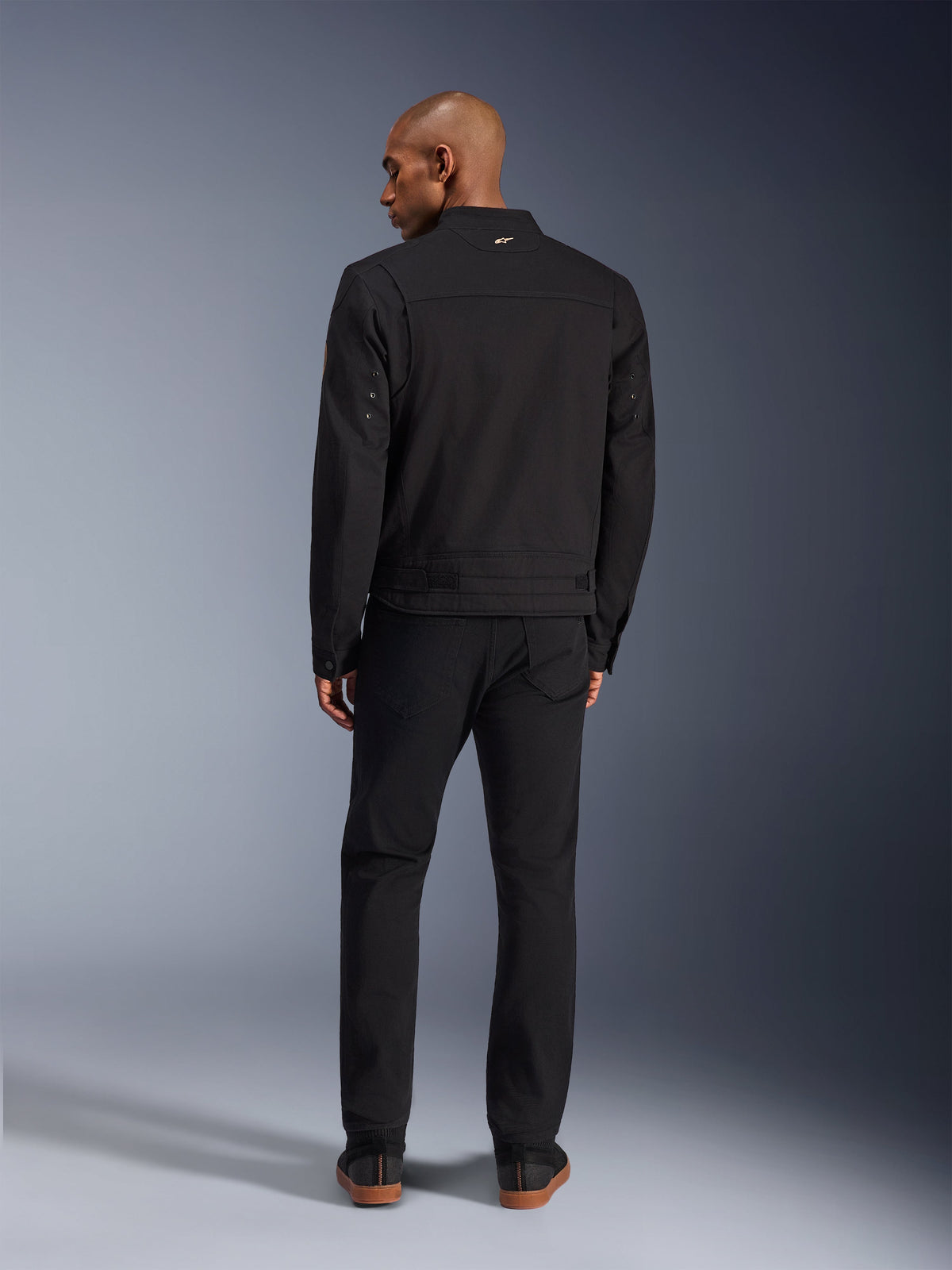 Chaqueta Alpinestars Bruiser en negro, vista posterior de modelo masculino con la chaqueta de moto urbana y pantalones negros a juego, con un pequeño logo dorado en el cuello y correas de ajuste en la cintura, estética inspirada en la ropa de trabajo