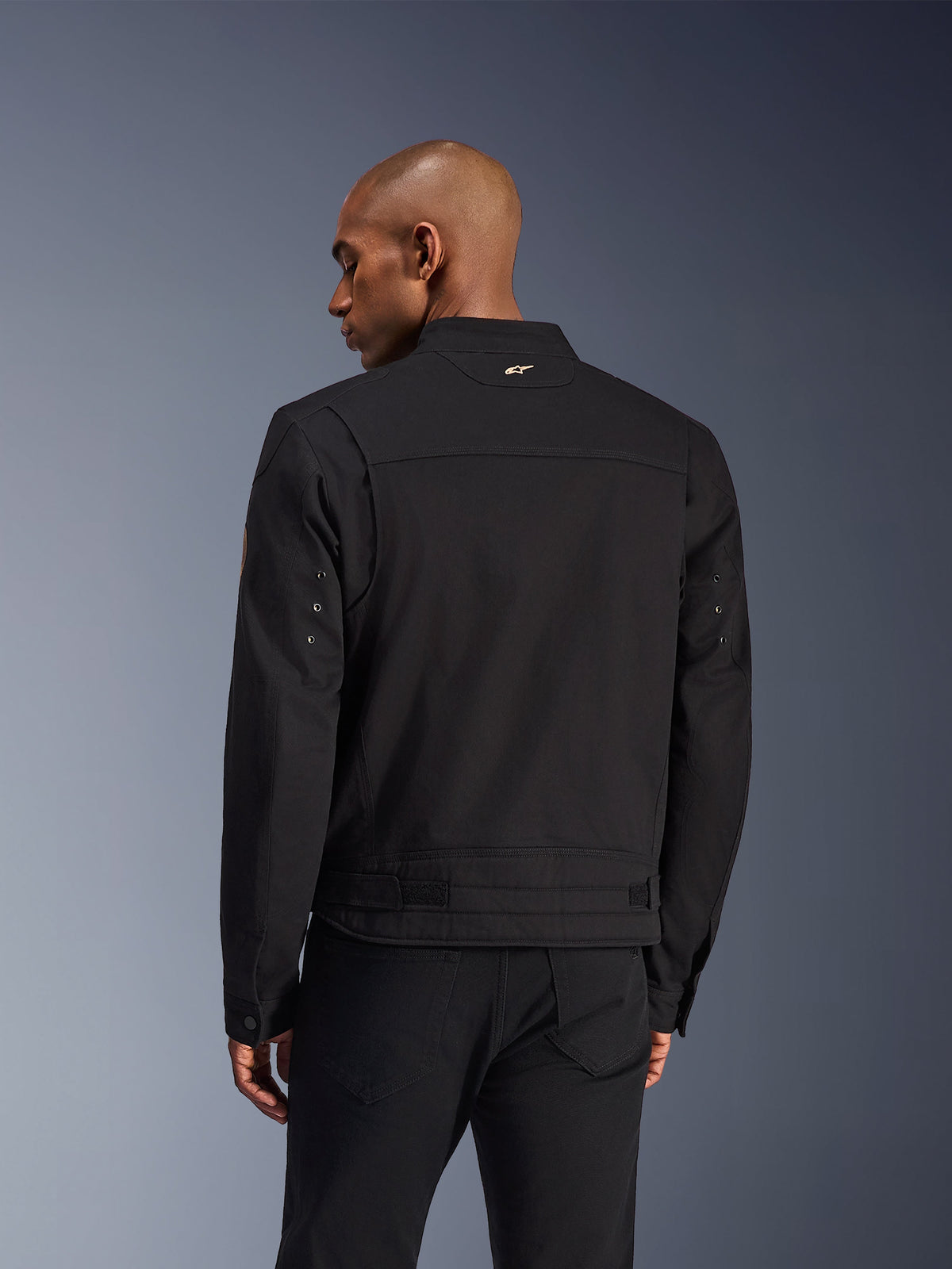 Chaqueta Alpinestars Bruiser, chaqueta de moto urbana negra, vista trasera, con un diseño inspirado en la ropa de trabajo, un pequeño logo dorado en el cuello y correas de ajuste en la cintura, construcción textil duradera para conducción urbana