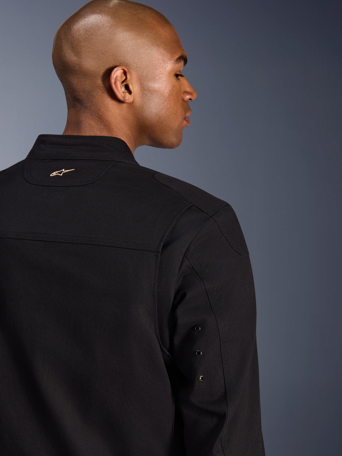 Alpinestars Bruiser Jacket, chaqueta textil urbana para motocicleta, negra, vista de cerca de la parte posterior con logotipo dorado bordado, detalles de ojales metálicos en la manga y paneles plisados en los hombros para una mayor movilidad