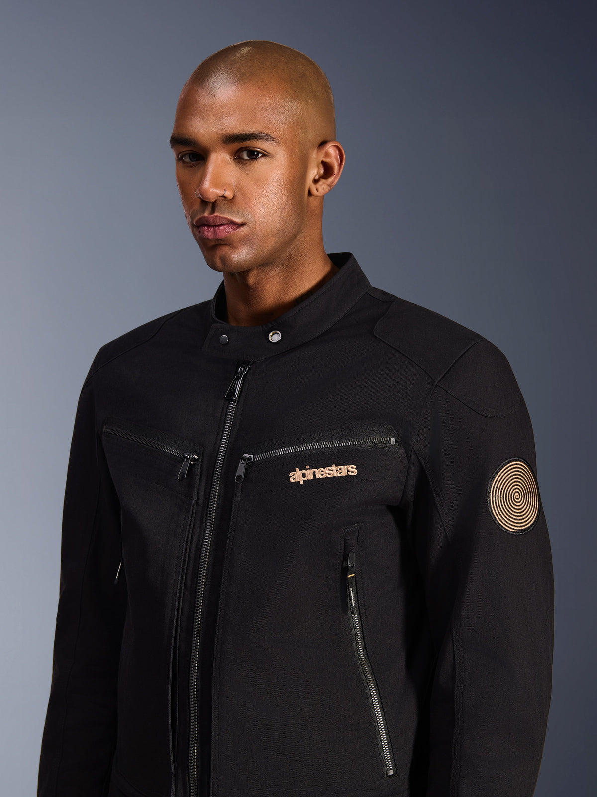 Modelo masculino con la Chaqueta Alpinestars Bruiser, negra, chaqueta de moto urbana en sarga de policotón, con bolsillos con cremallera, logotipo bordado en dorado en el pecho y parche espiral Oscar en la manga, estilo urbano moderno