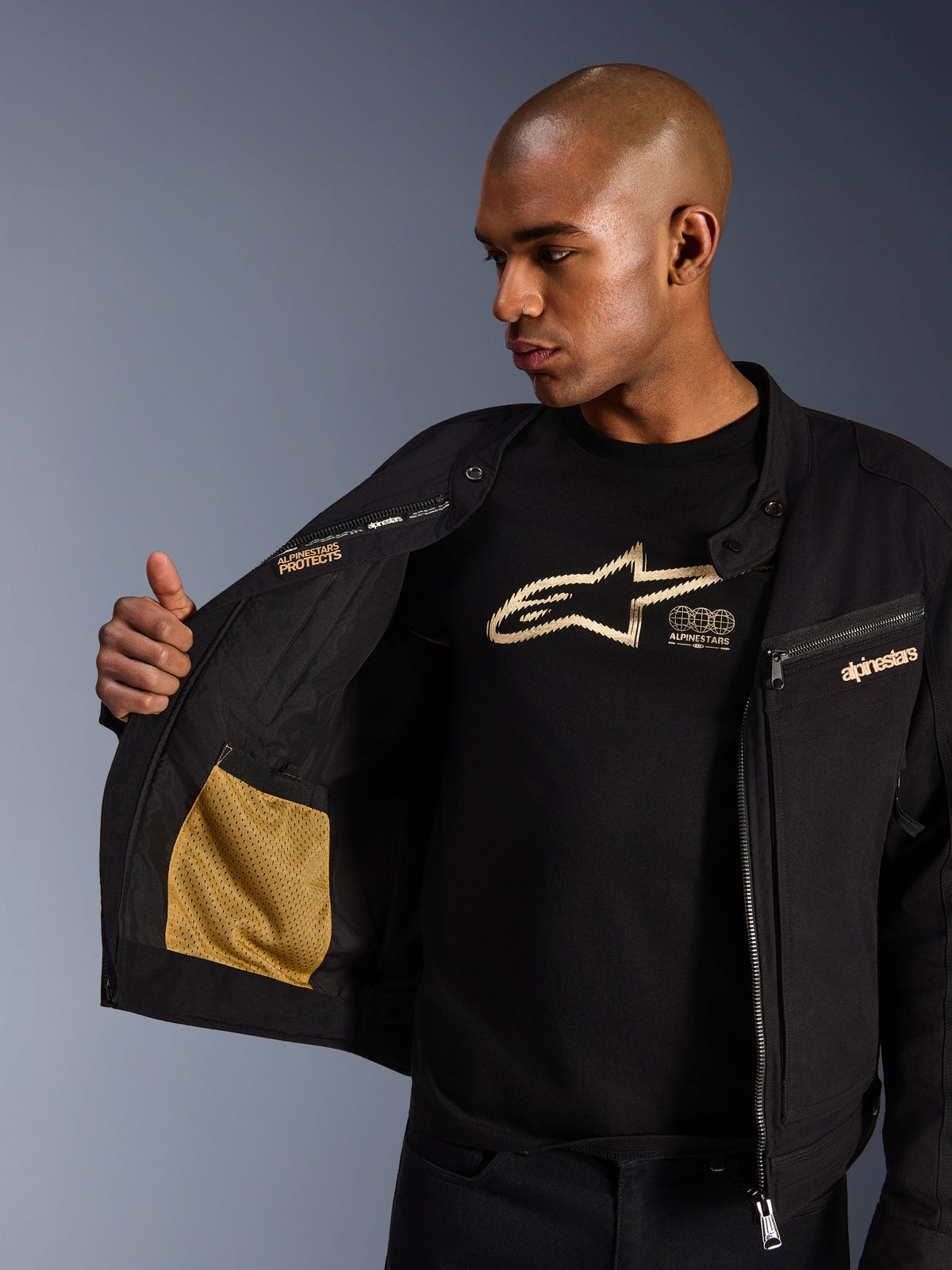 Chaqueta Alpinestars Bruiser, chaqueta urbana de motociclismo en color negro, vista de primer plano que muestra el interior abierto con bolsillo de malla dorada y marca 'Alpinestars Protects', usada sobre una camiseta negra con logotipo, equipamiento textil de conducción con estética de ropa de trabajo