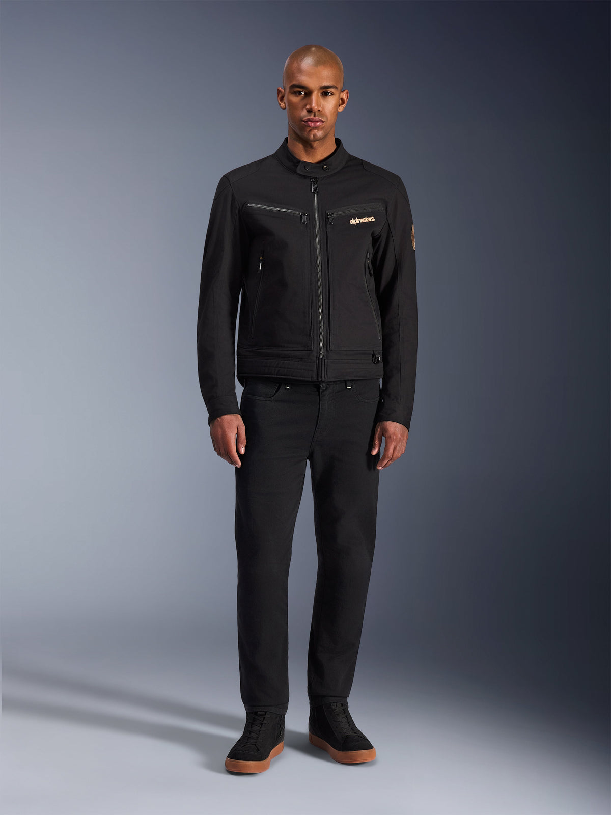 Chaqueta Alpinestars Bruiser, chaqueta de moto urbana negra, usada por un modelo con pantalones negros y zapatillas de caña alta, vista frontal que muestra los bolsillos del pecho con cremallera y el logotipo bordado, diseño inspirado en la ropa de trabajo para trayectos diarios
