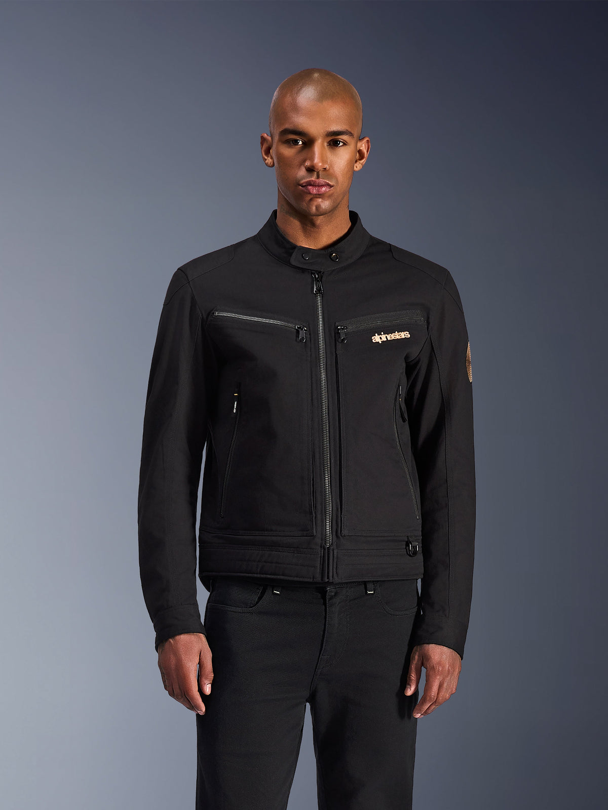 Chaqueta Alpinestars Bruiser, chaqueta de moto urbana negra, vista frontal en un modelo, con una estética clásica de ropa de trabajo, bolsillos con cremallera en el pecho, cuello con botón y logotipo de Alpinestars, diseñada para motociclistas urbanos