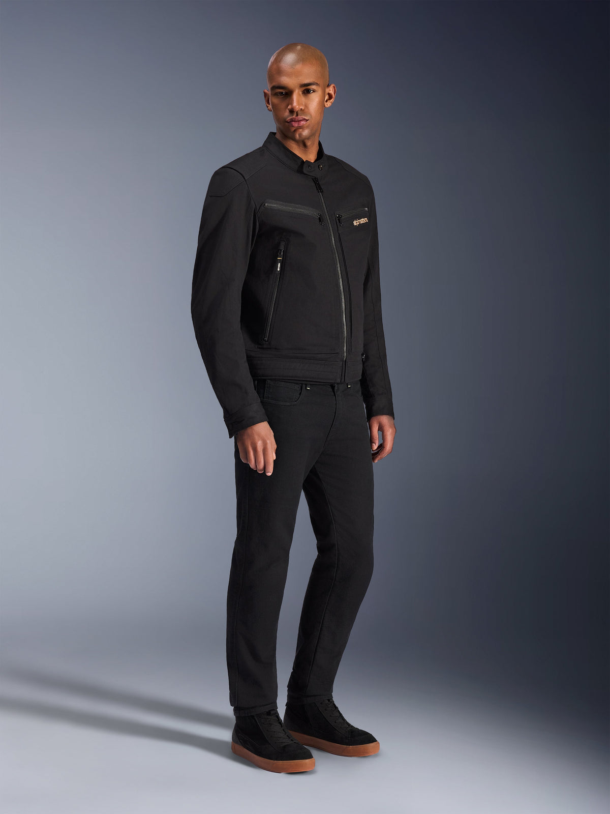 Chaqueta Alpinestars Bruiser, chaqueta de moto urbana, negra, lucida por un modelo con pantalones negros y zapatillas, con bolsillos en el pecho con cremallera, tejido de sarga de policotón y un diseño elegante inspirado en la ropa de trabajo para la conducción urbana