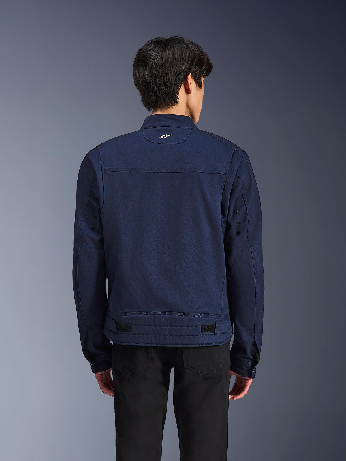 Chaqueta Alpinestars Bruiser, chaqueta de moto textil en azul oscuro, vista posterior en modelo, diseño inspirado en la ropa de trabajo con un pequeño logo blanco de Astars en el cuello, pestañas de ajuste en la cintura, ropa de conducción urbana contemporánea.